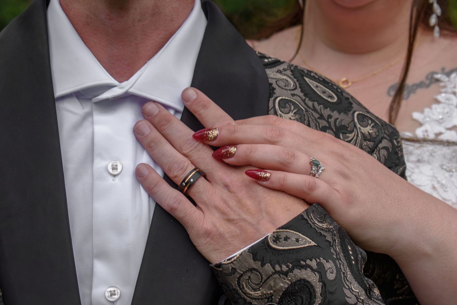 Close-up of a married couple's hands showing wedding rings, with red manicured nails and a gray suit jacket.