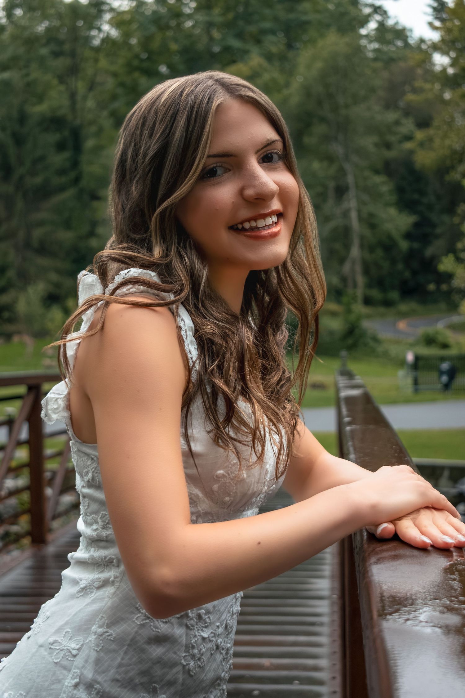 Young woman in white lace dress smiling on wooden bridge surrounded by lush green trees.