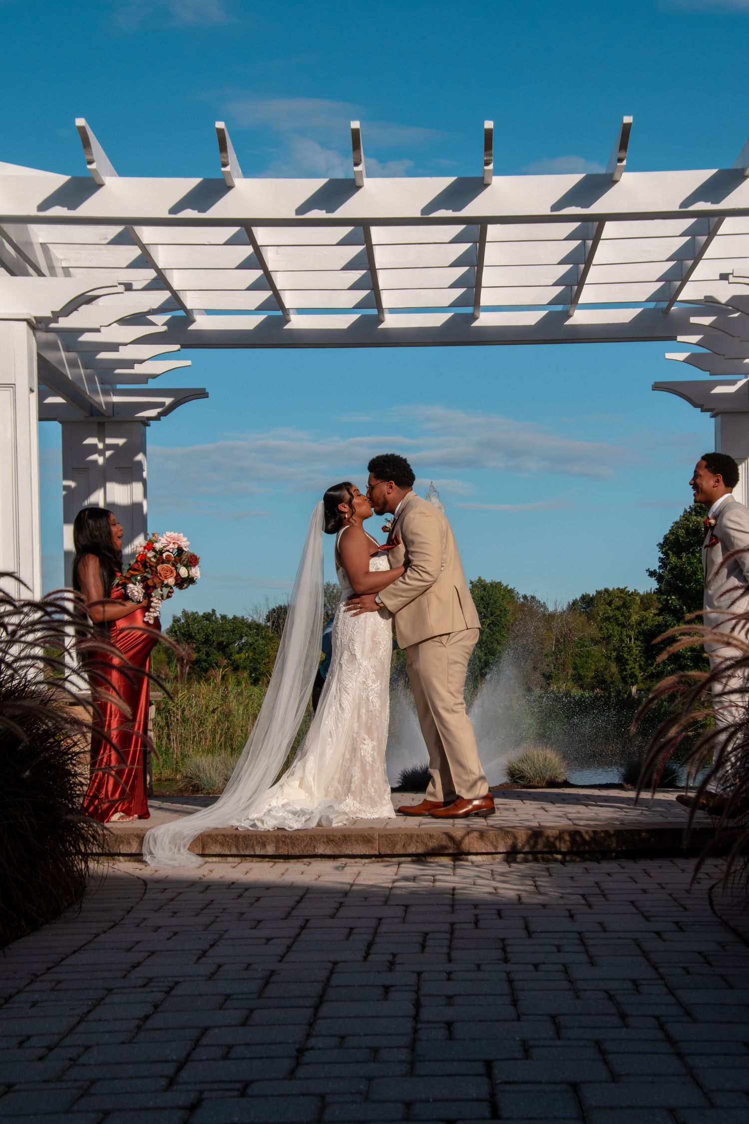 Bride and groom share first kiss under white pergola with fountain backdrop on sunny wedding day.