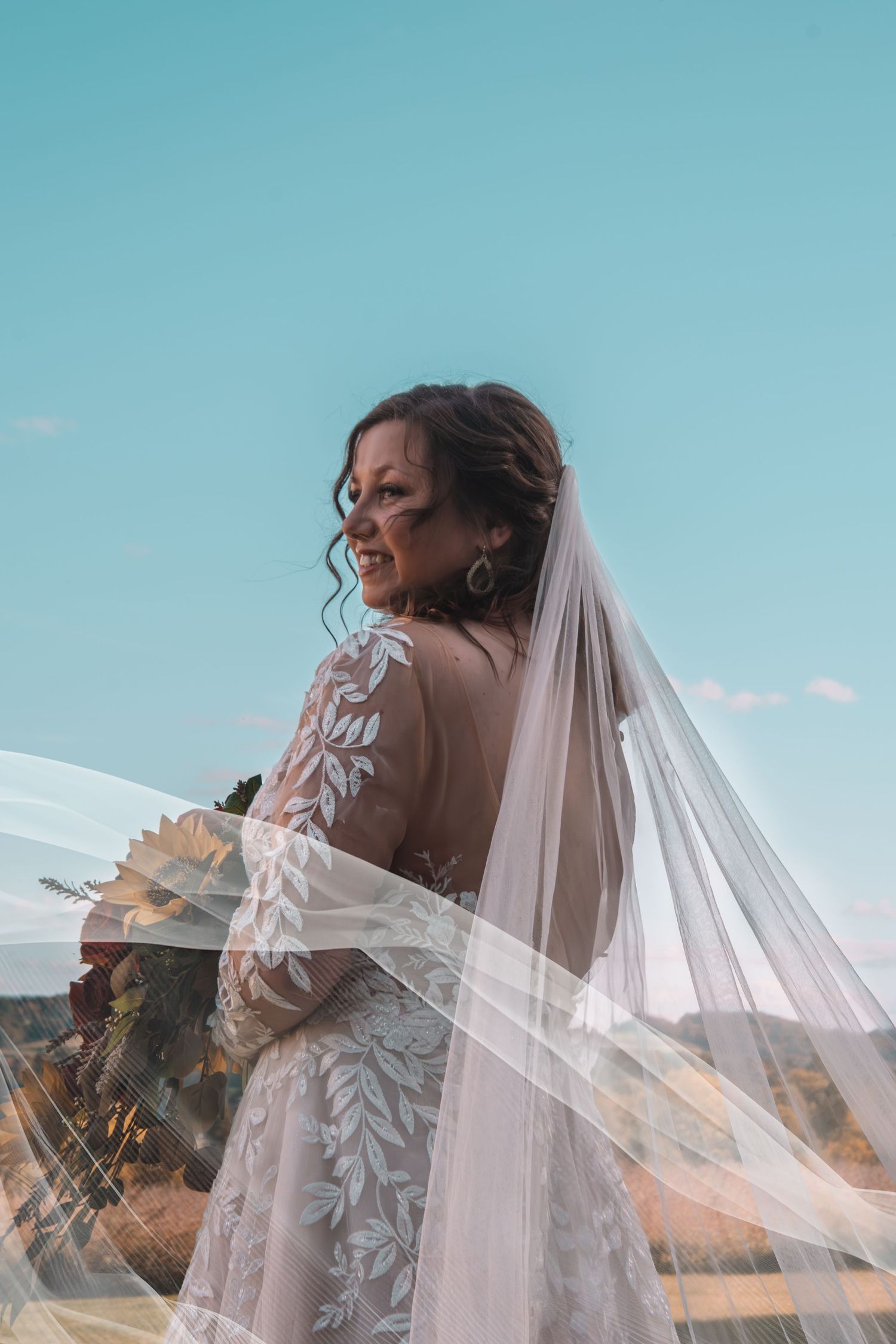 Smiling bride in lace long-sleeve gown with flowing veil against blue sky outdoors.