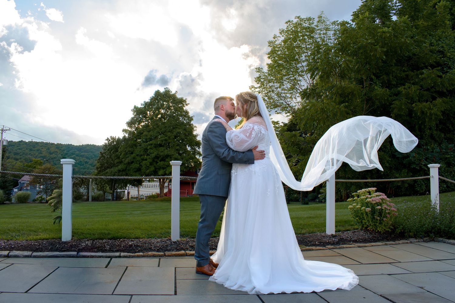 Bride and groom share a romantic kiss outdoors, with her white veil flowing in the wind against a green landscape. The Barn at Wight Farm