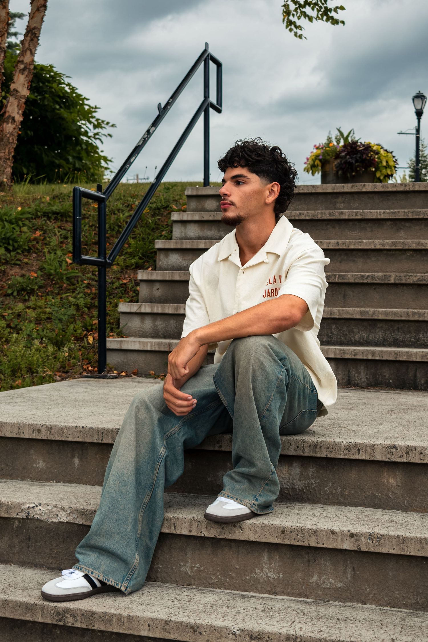 Young man in white shirt and wide-leg jeans sitting on concrete steps outdoors, looking thoughtfully to the side.