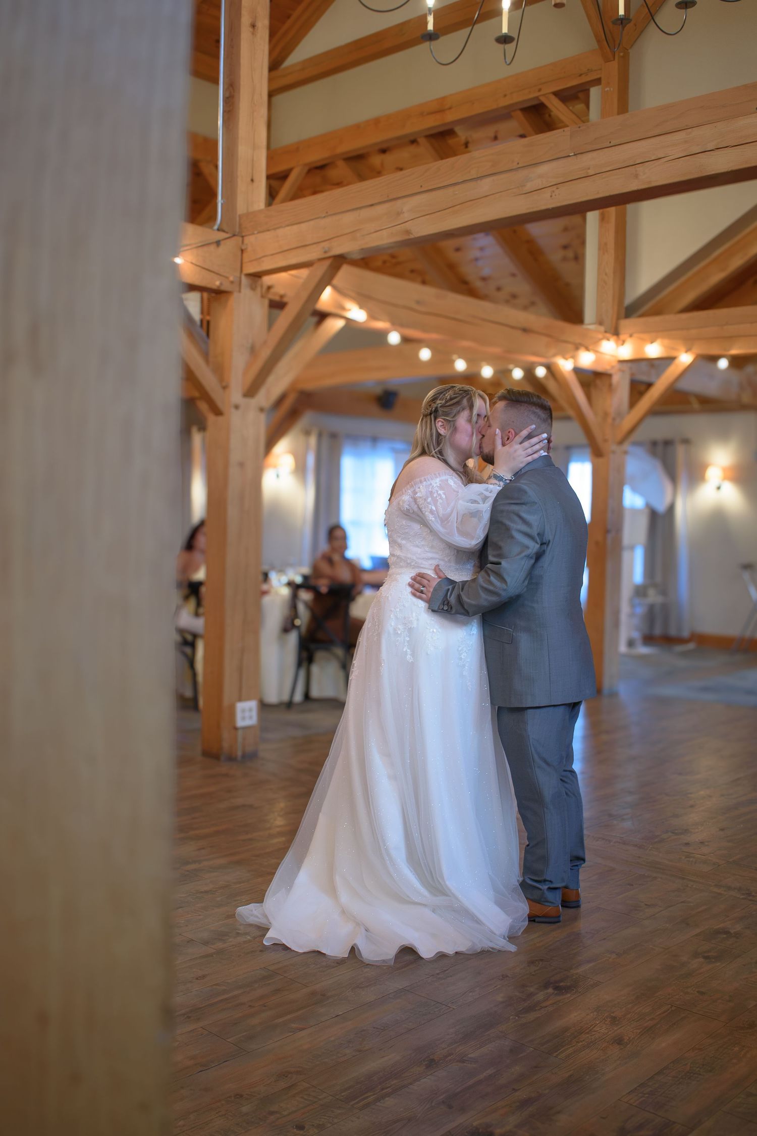 Bride and groom share first dance kiss in rustic barn venue with wooden beams and string lights. Massachusetts, Sturbridge Village