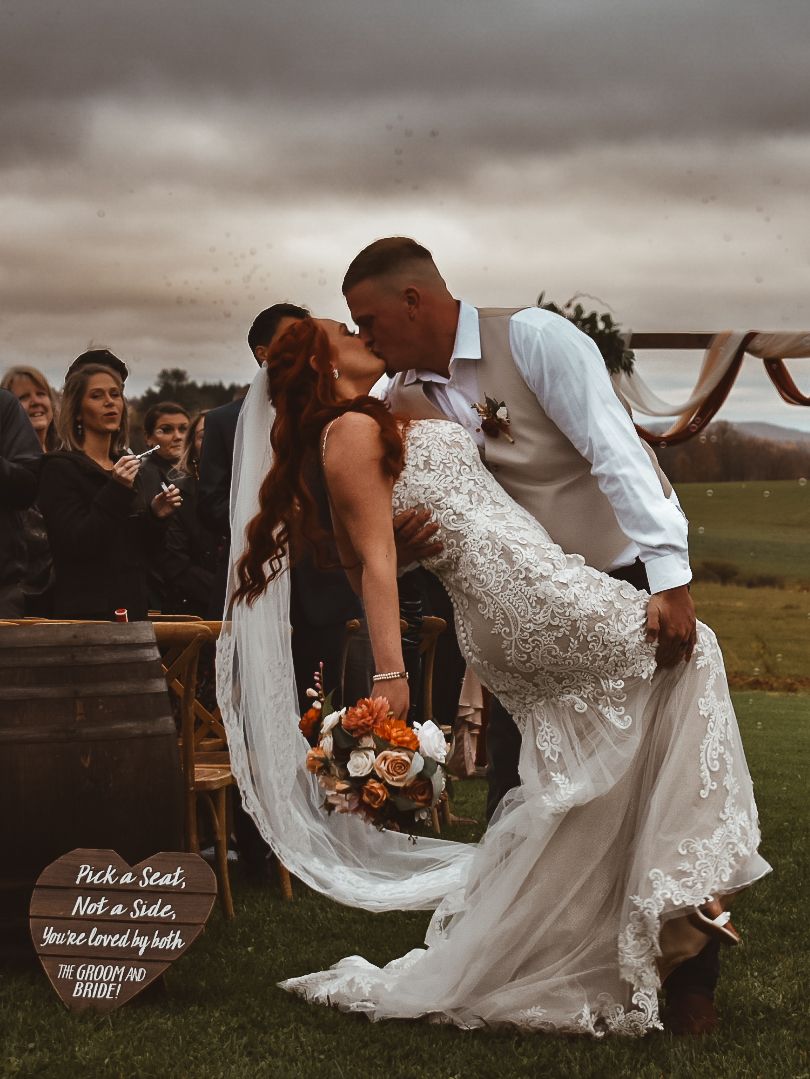 Bride and groom share a dramatic dip kiss at outdoor wedding ceremony under cloudy skies.