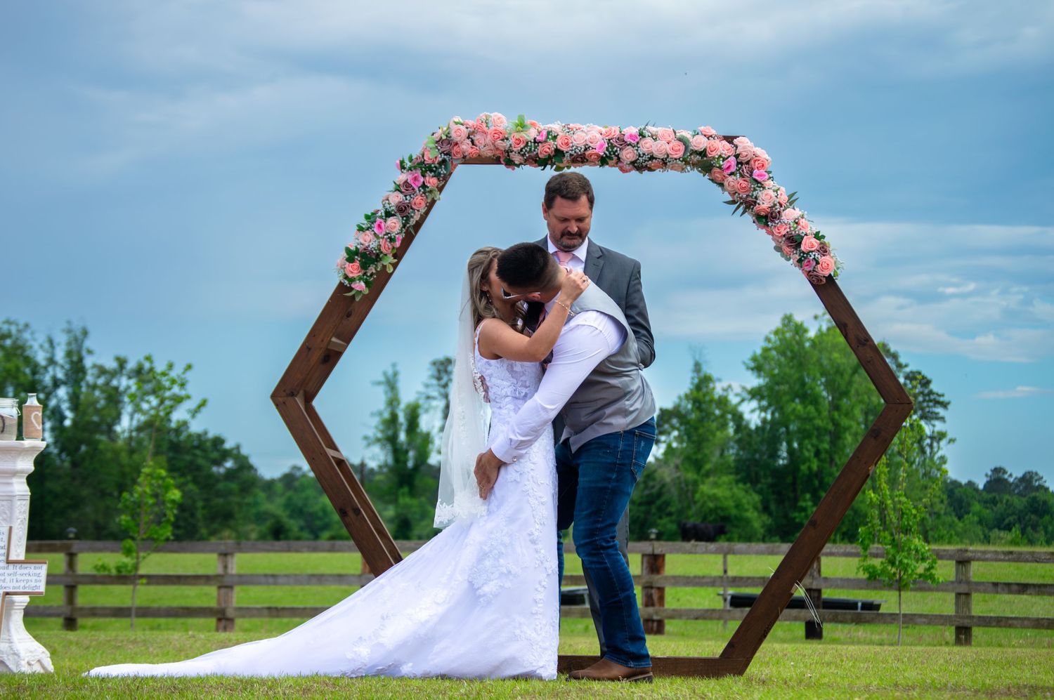 Bride and groom share a kiss under a hexagonal wooden arch adorned with pink flowers at an outdoor wedding ceremony.
