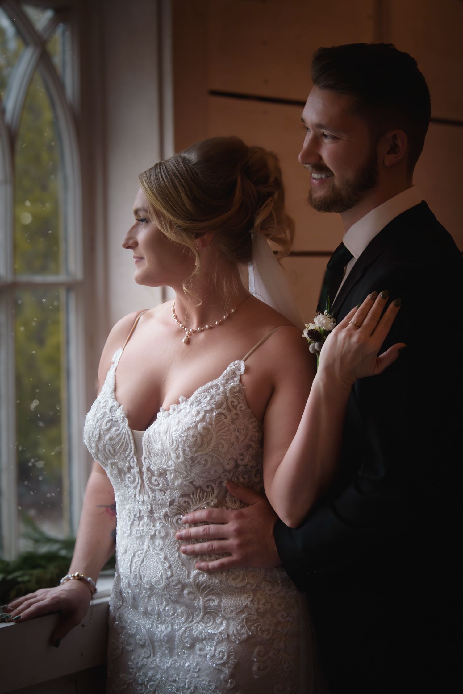 Bride in lace gown and groom in dark suit stand together gazing out a rain-streaked window in soft natural light.