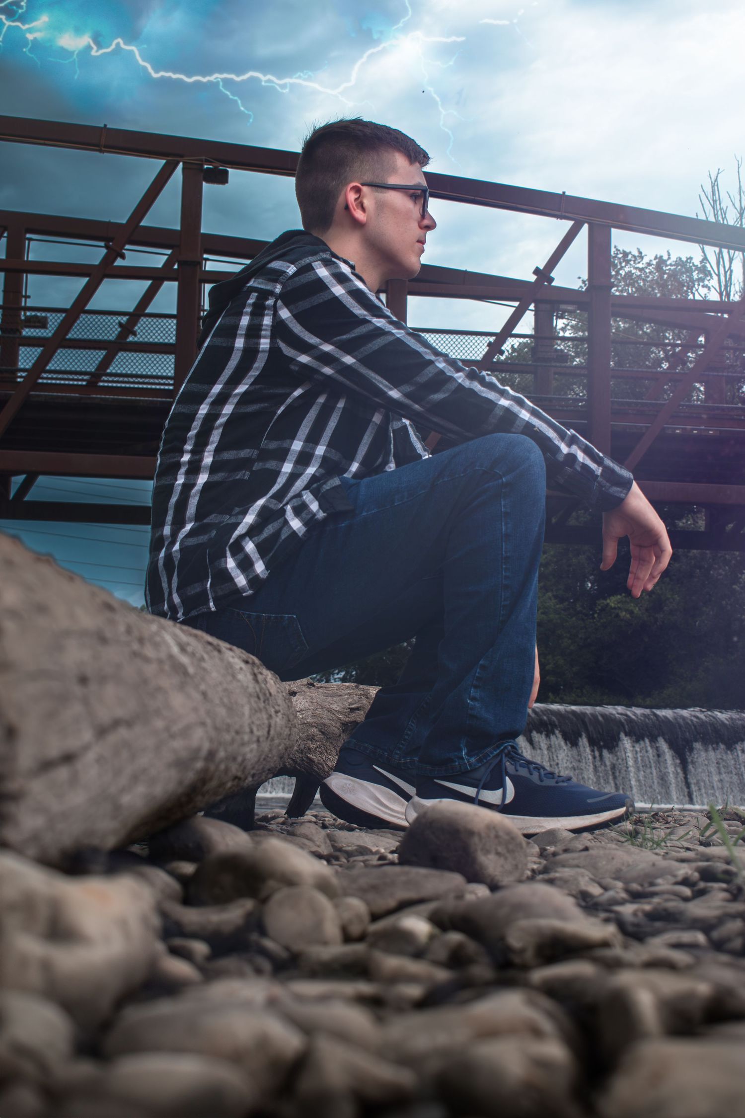 Young man in plaid shirt and jeans crouching on rocky riverbank near a metal bridge under stormy sky.