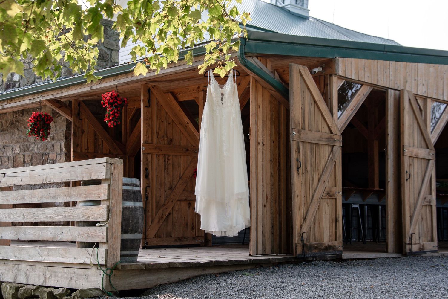 White wedding dress hanging outside rustic wooden barn doors with red floral decorations.