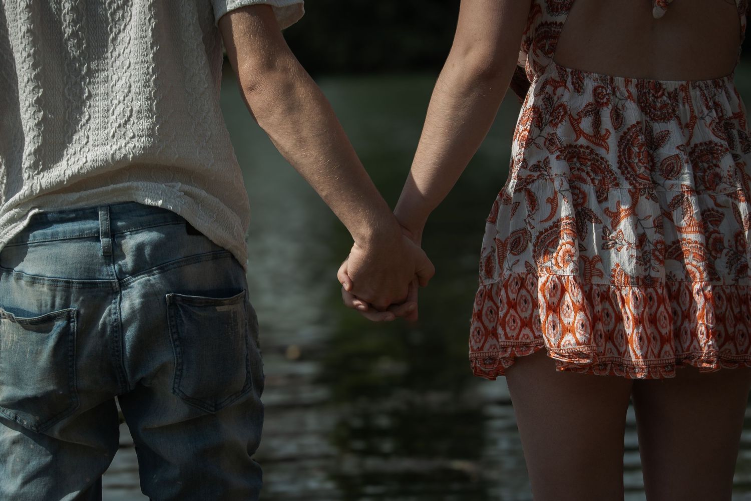 Couple holding hands near water, wearing casual summer clothes, romantic outdoor moment.