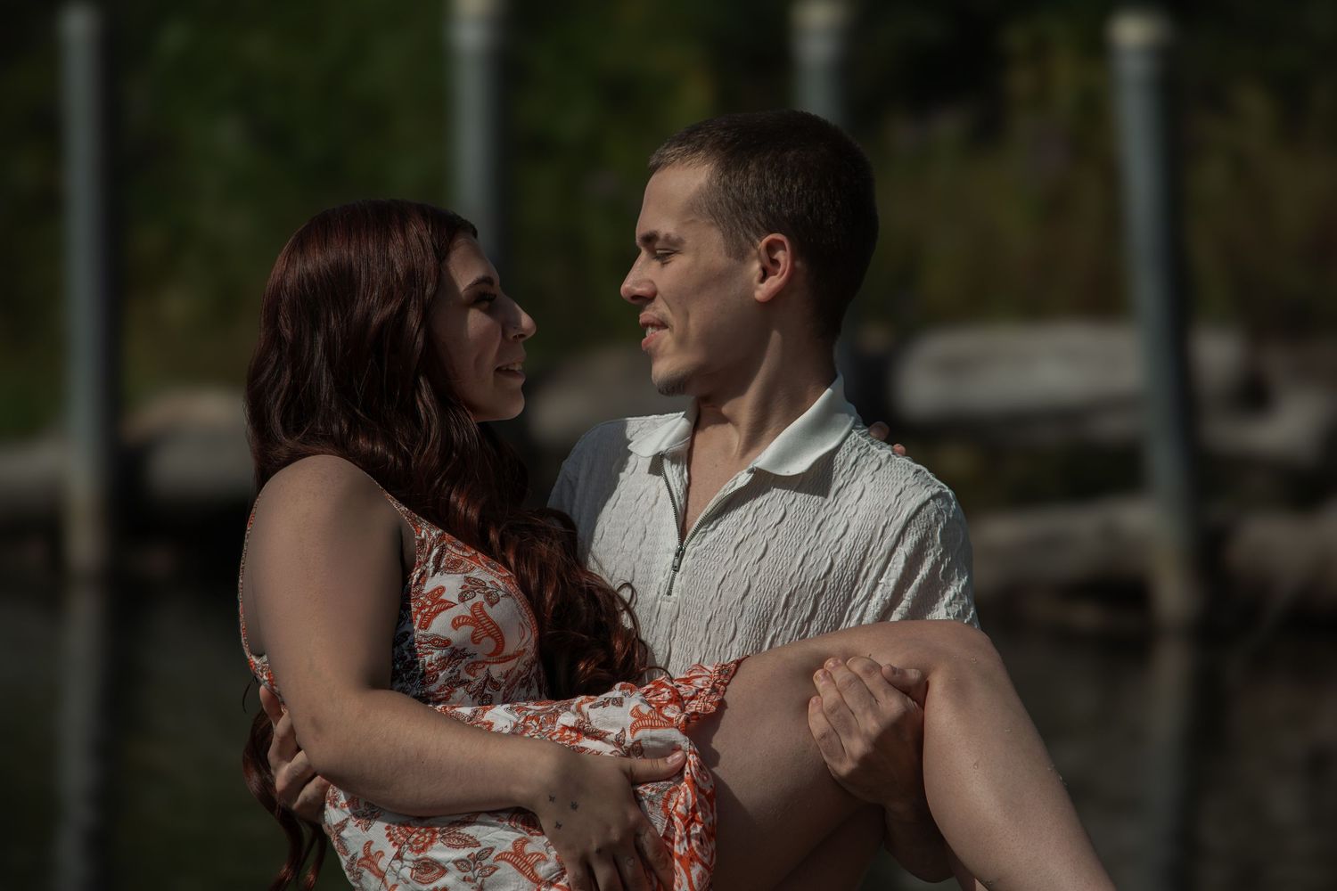 A couple embraces romantically near a waterfront dock, the woman in a floral dress and man in a white shirt gazing at each other.