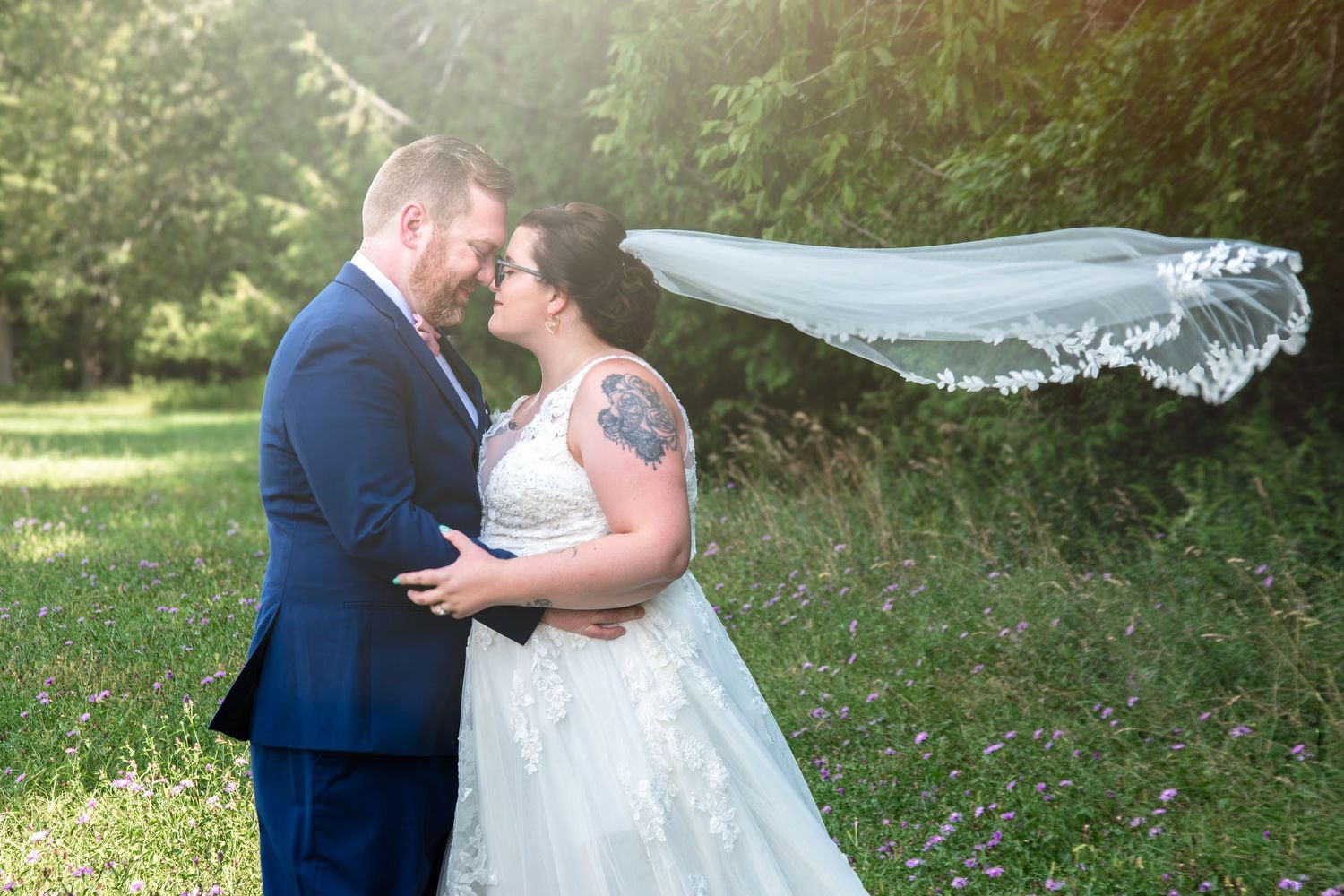 Bride and groom share romantic moment outdoors, veil flowing in wind, surrounded by lush greenery in soft sunlight.