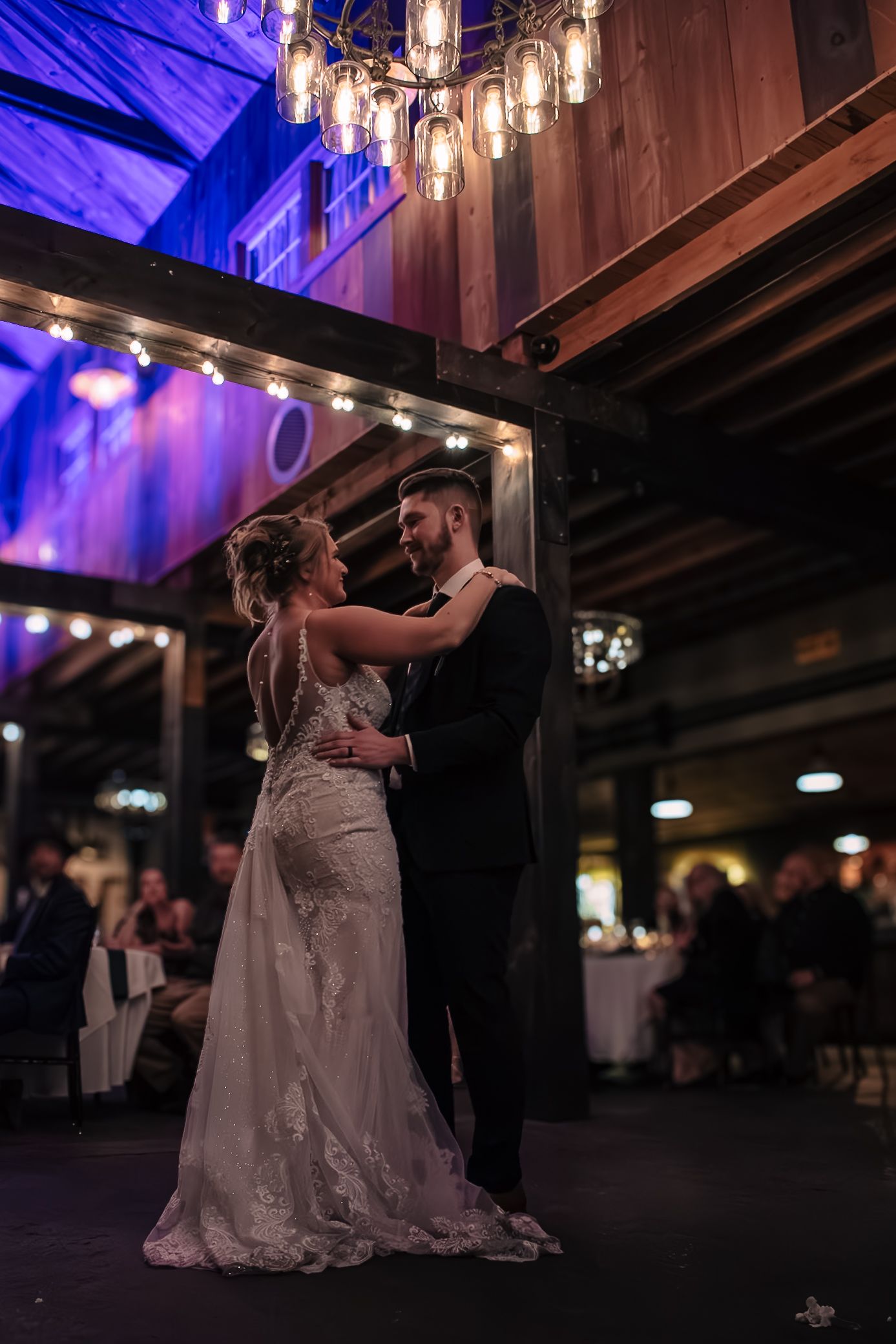 Bride and groom share their first dance under glowing Edison bulb chandelier in a rustic wedding reception venue.