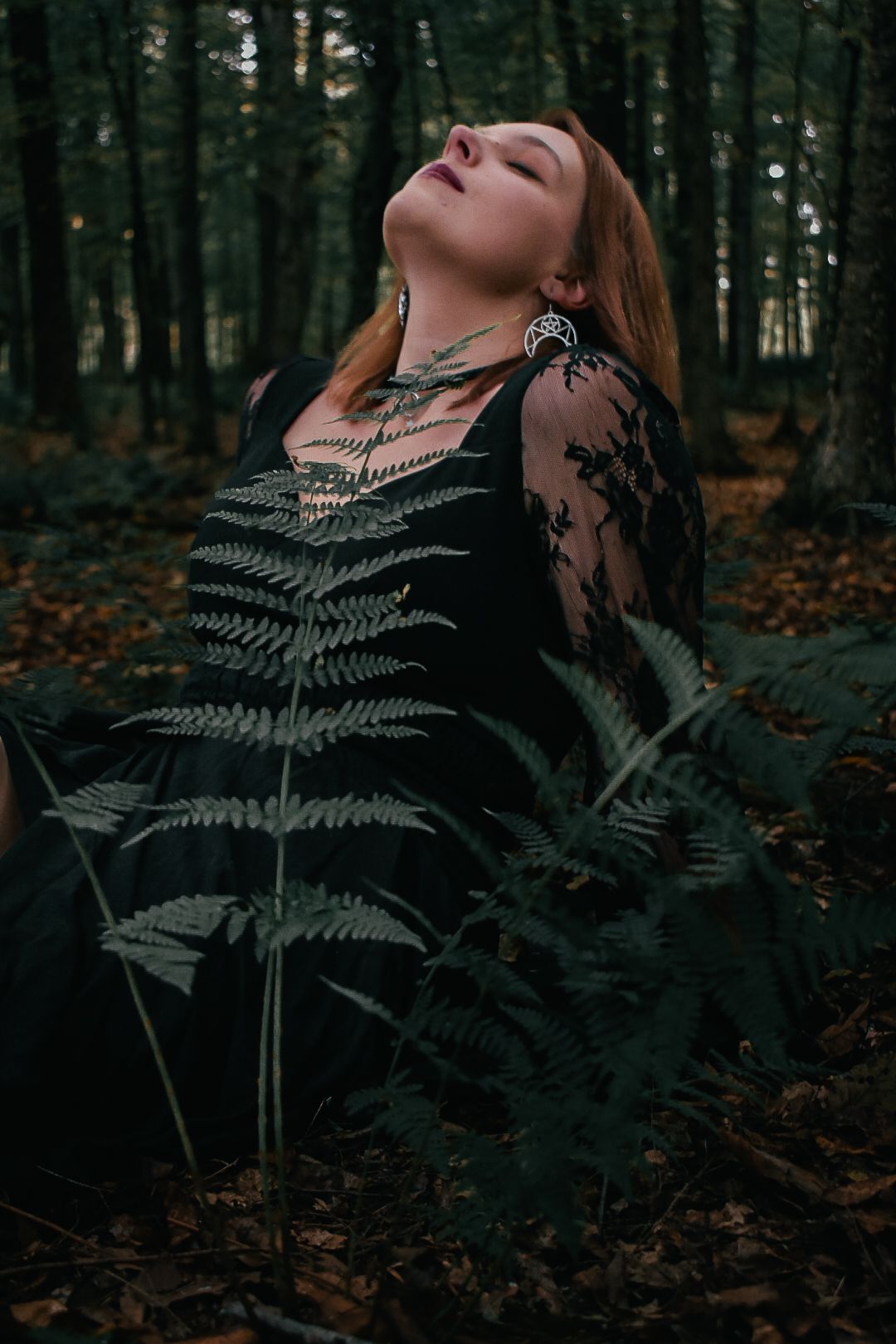 Redhead woman in black lace dress sitting among ferns in dark forest with eyes closed.