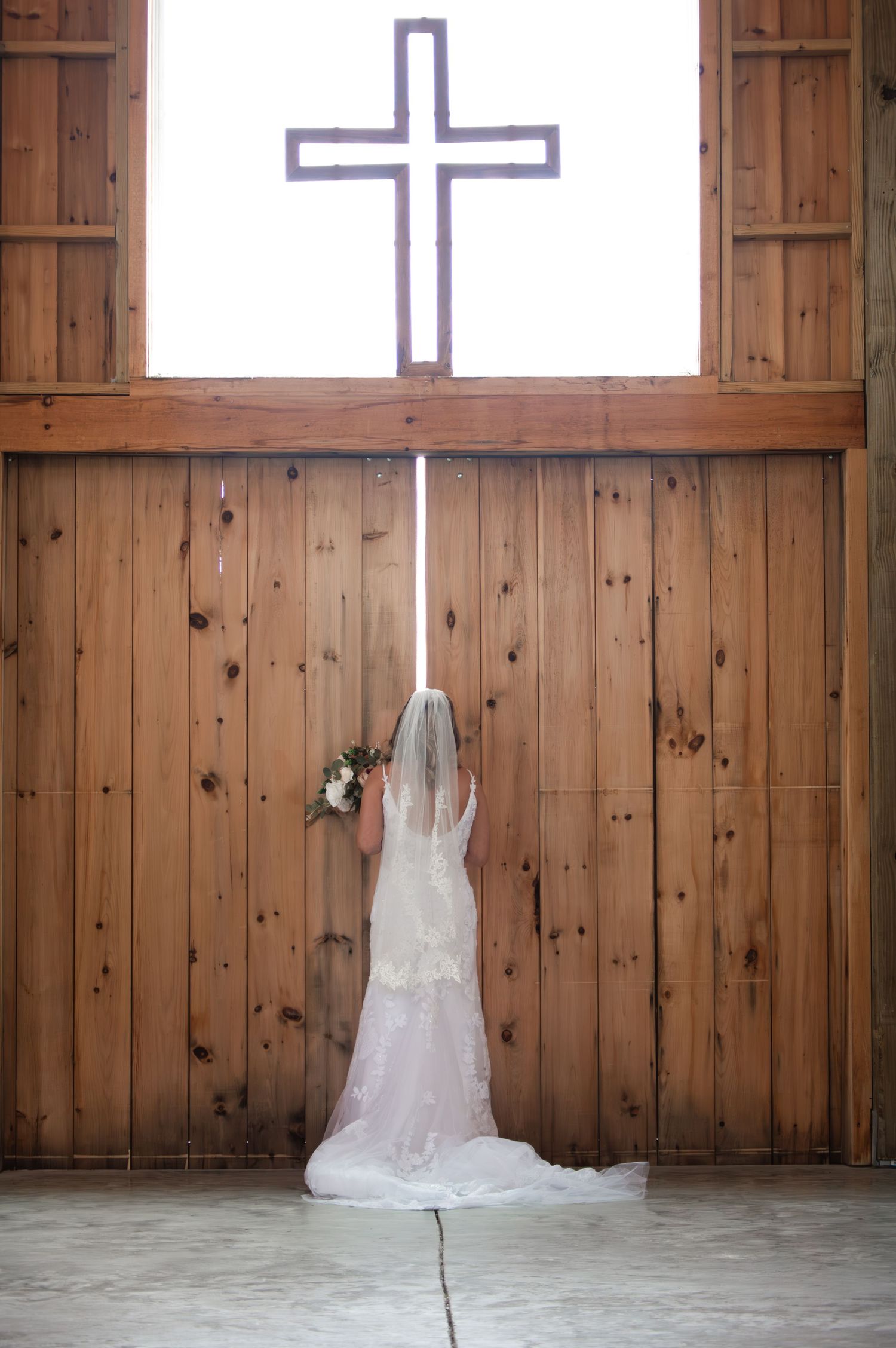 Bride in lace wedding dress and veil holding bouquet stands before wooden chapel doors with cross.