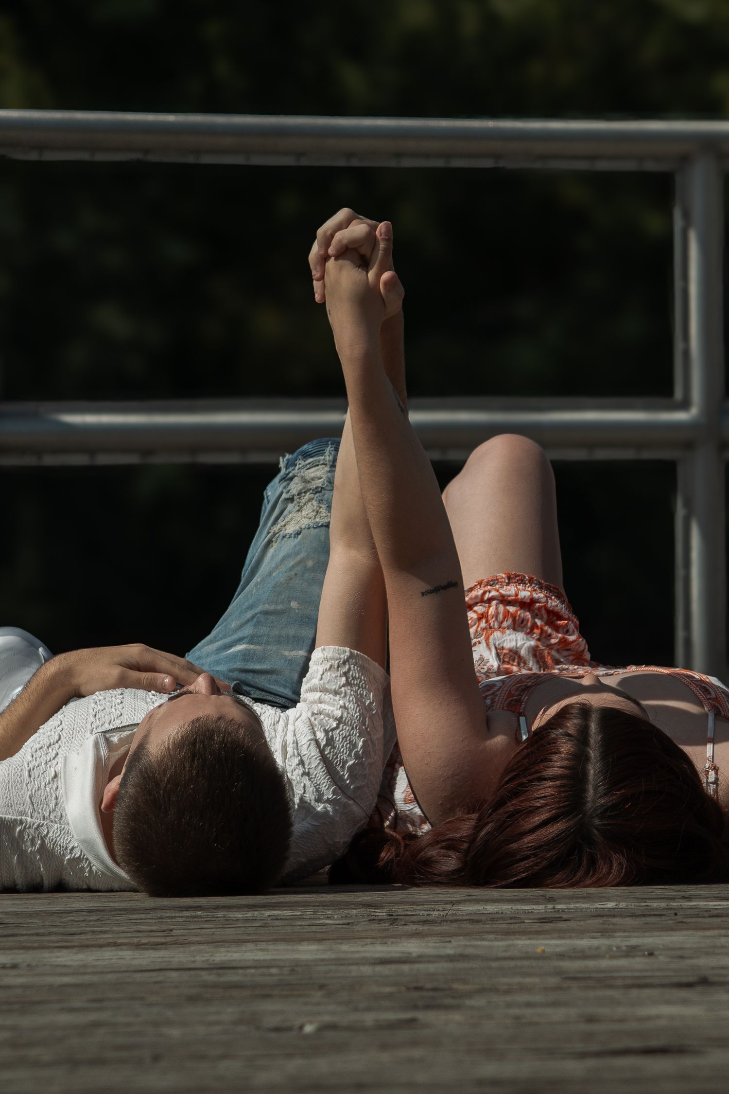 Couple lying on wooden dock holding hands raised in air on sunny day with green trees in background.