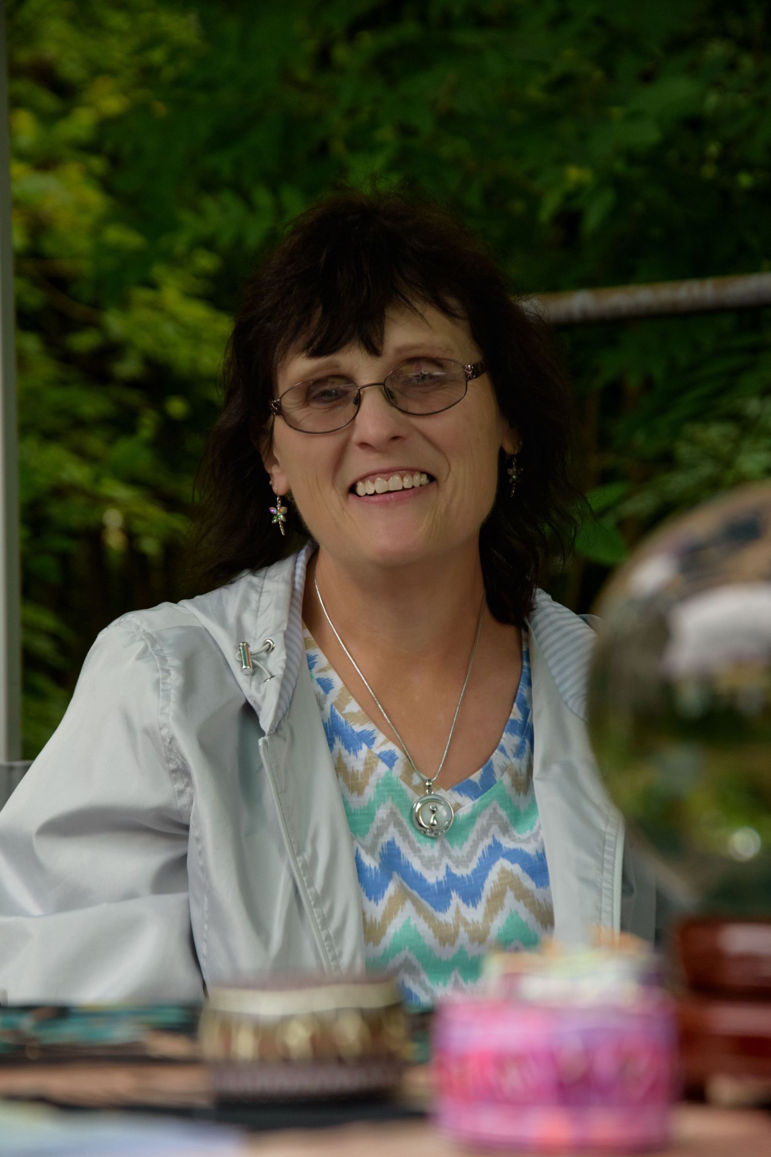 Smiling middle-aged woman with dark hair and glasses wearing a colorful zigzag top outdoors.