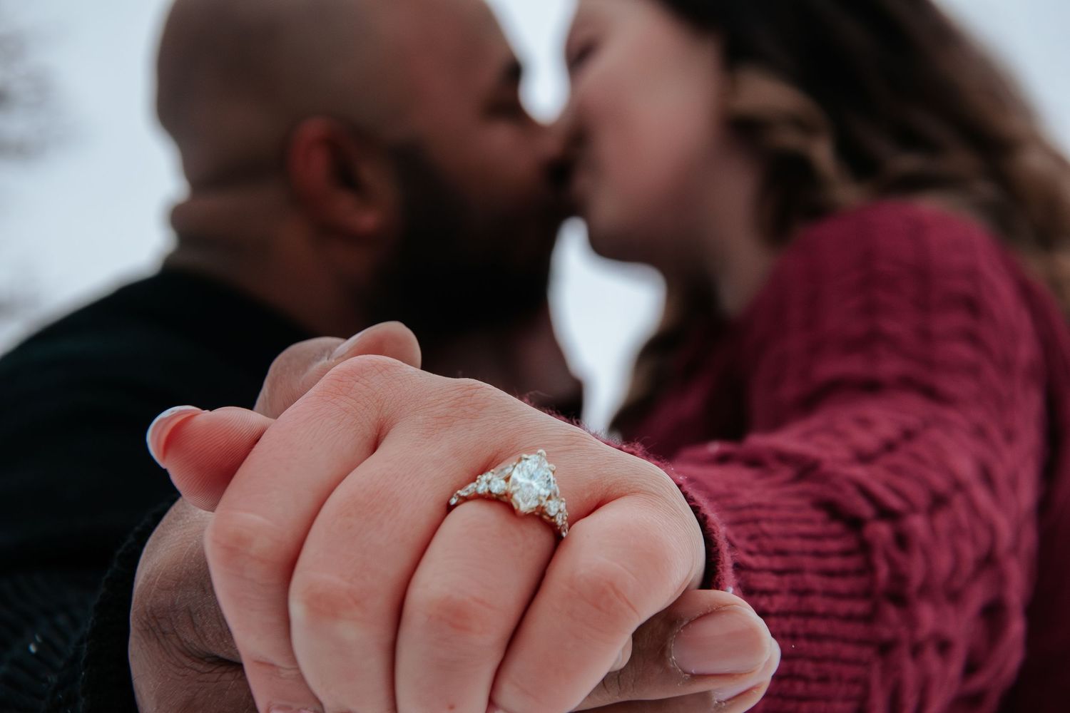 Couple kissing in winter while showcasing a large oval diamond engagement ring on a gold band.
