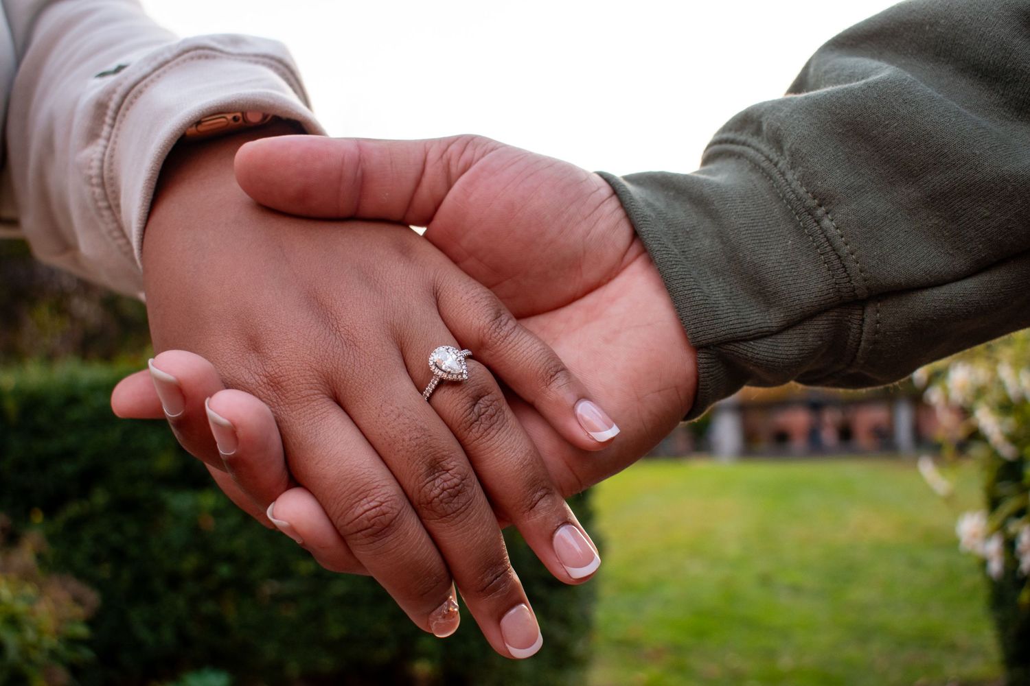 Two people holding hands outdoors, one wearing a diamond engagement ring, symbolizing love and commitment.