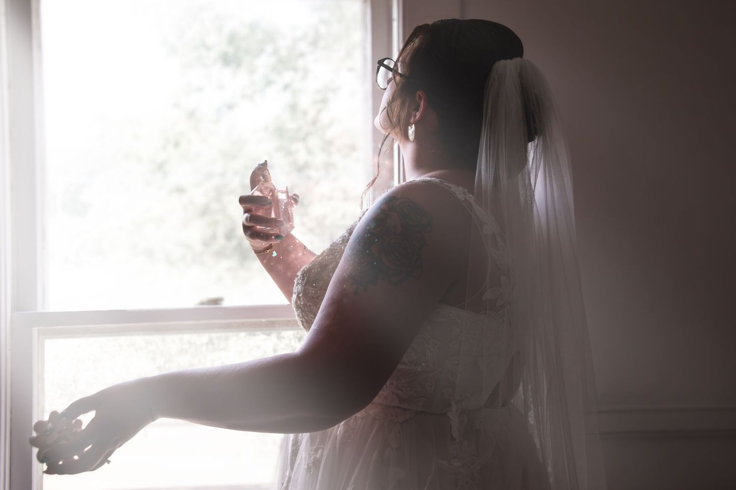 Silhouette of a bride in wedding dress and veil holding perfume bottle by a bright window.