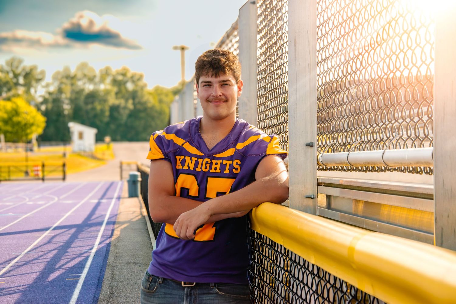 Teen boy in purple Knights football jersey leaning against a fence at a high school track field during golden hour.
