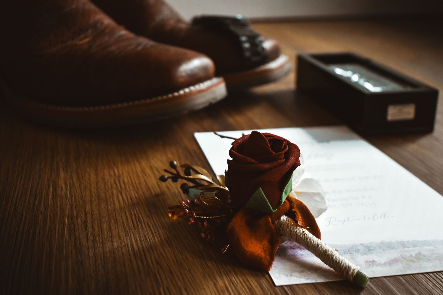 Dark red rose boutonniere with a handwritten letter beside brown leather boots on a wooden floor.