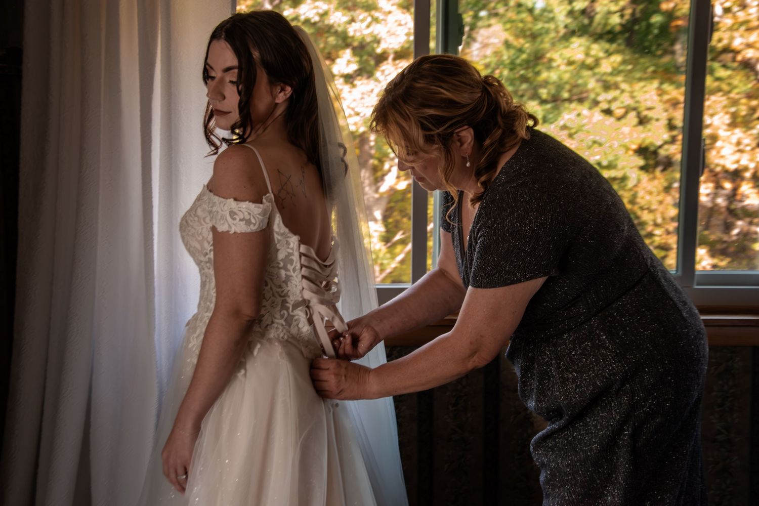 A bride in a white lace wedding dress and veil gets her corset back fastened by a woman near a window.