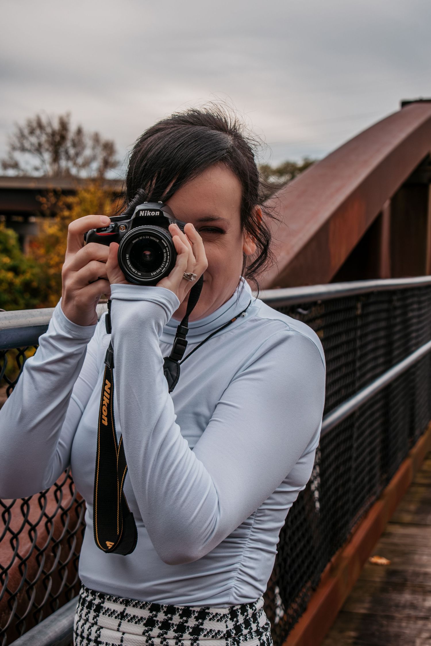Woman photographer holding a Nikon DSLR camera on a bridge outdoors on a cloudy day.