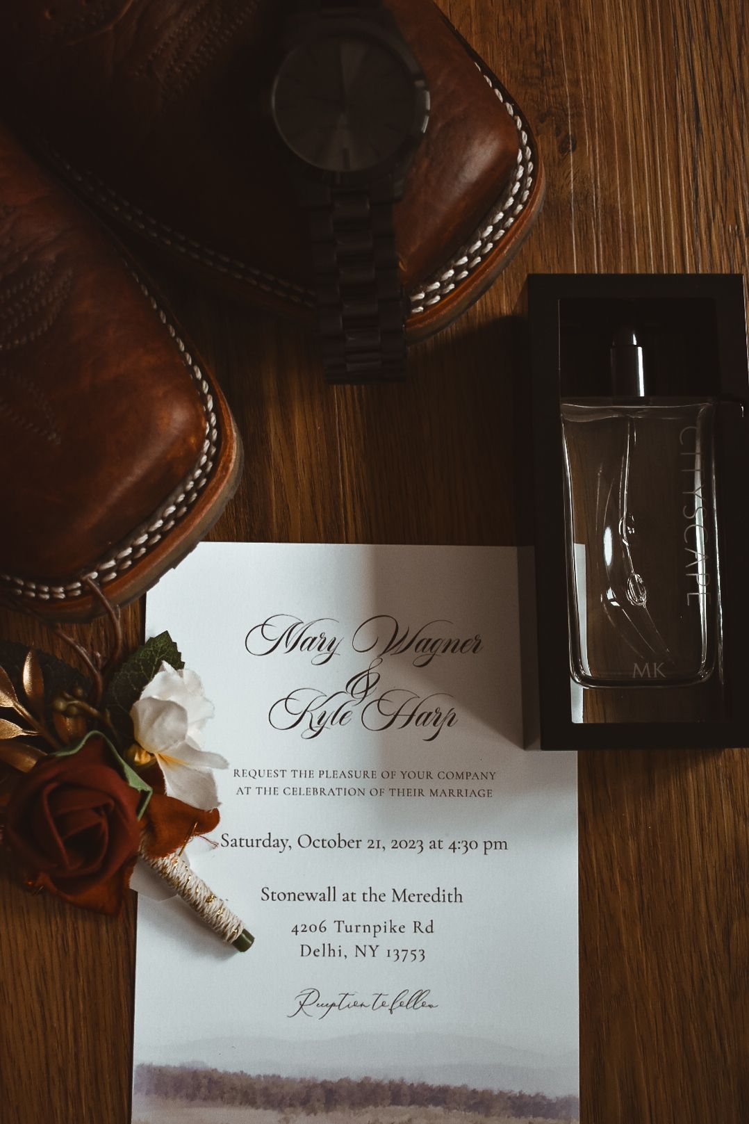 Wedding invitation for Mary Wagner and Kyle Hay beside leather boots, watch, and cologne.