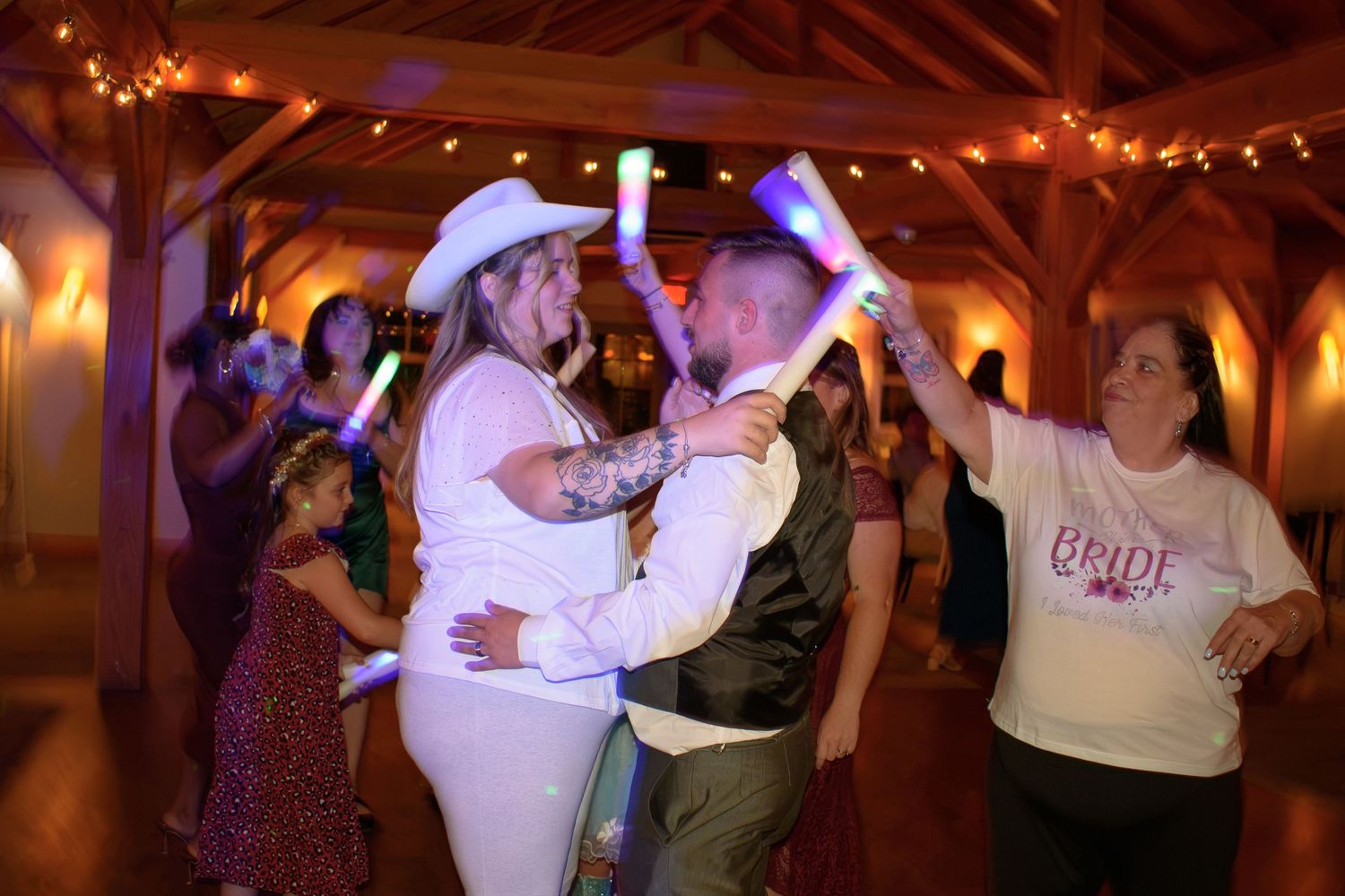 Bride and groom share first dance surrounded by guests holding glowing foam sticks at rustic barn wedding reception.