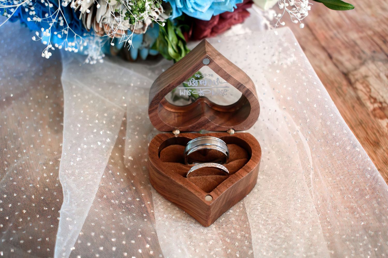 Two silver wedding bands displayed in a heart-shaped wooden ring box on white tulle fabric near a floral bouquet.