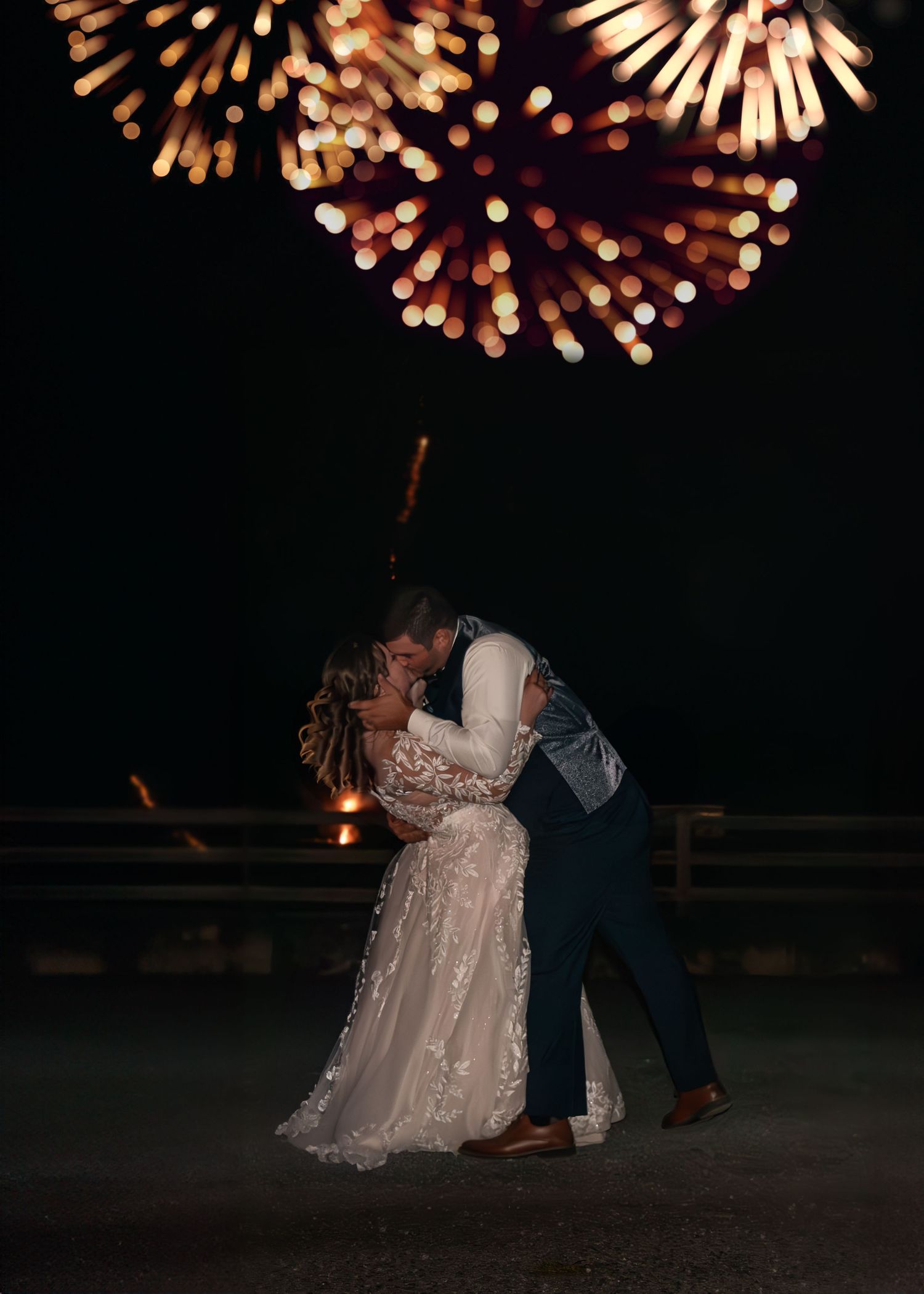 Bride and groom share a romantic kiss under glowing fireworks at night, with the bride in a lace gown.