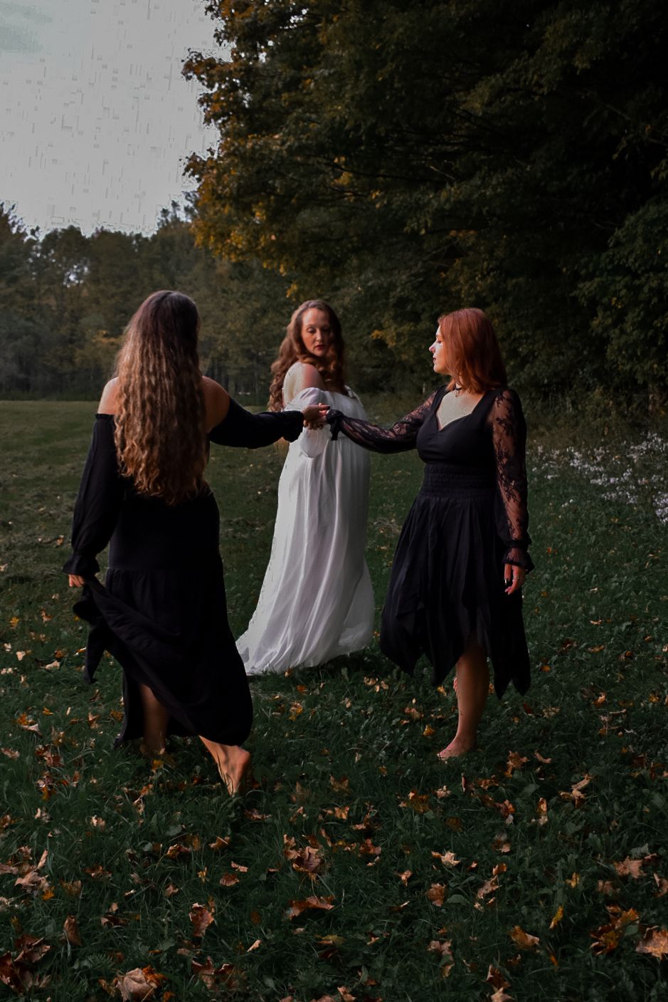 Three women in white and black dresses holding hands in a misty autumn forest clearing at dusk.