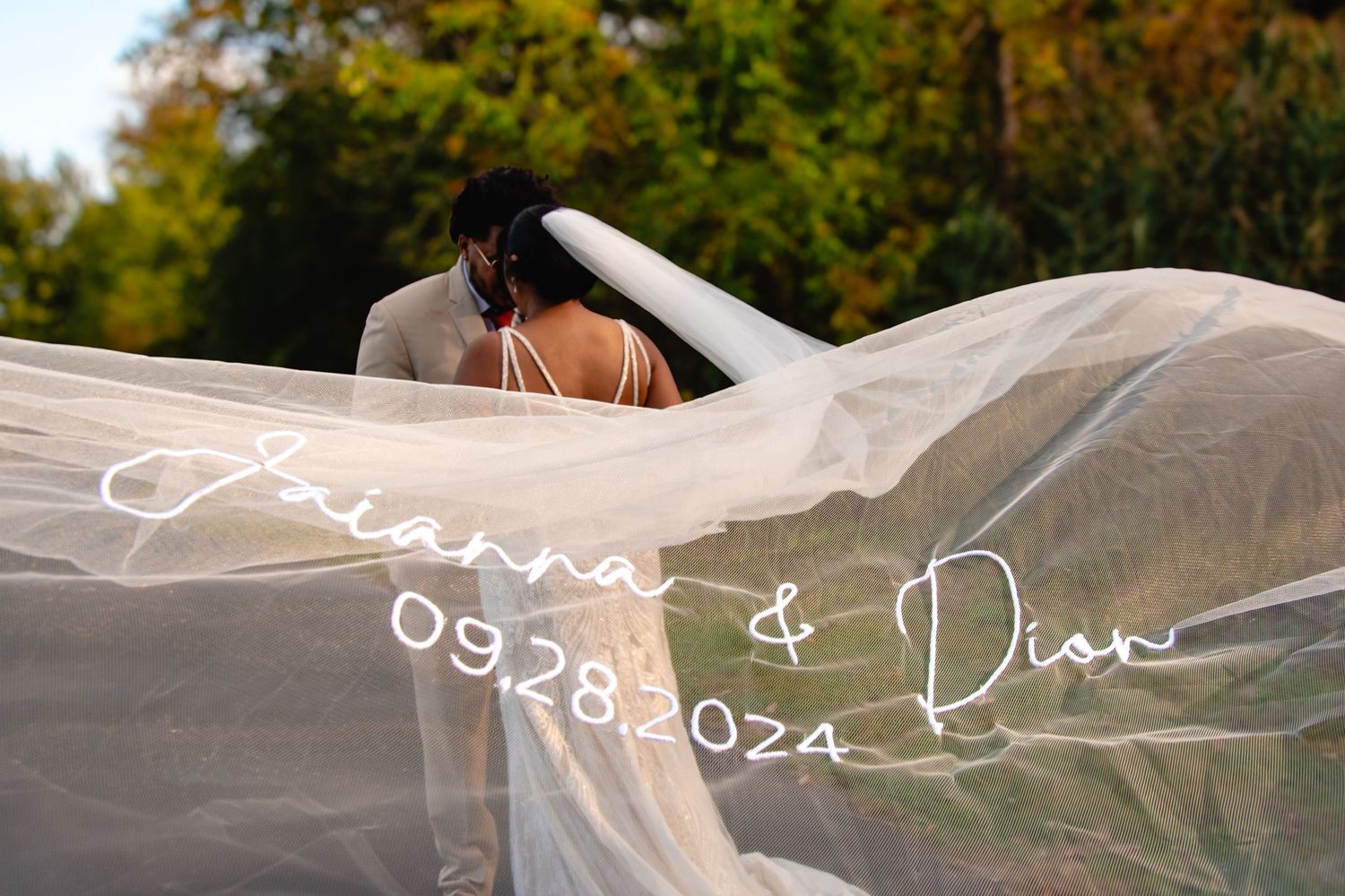 Bride and groom share a romantic kiss beneath a flowing white veil outdoors, with text reading Arianna & Dion 09.28.2024.
