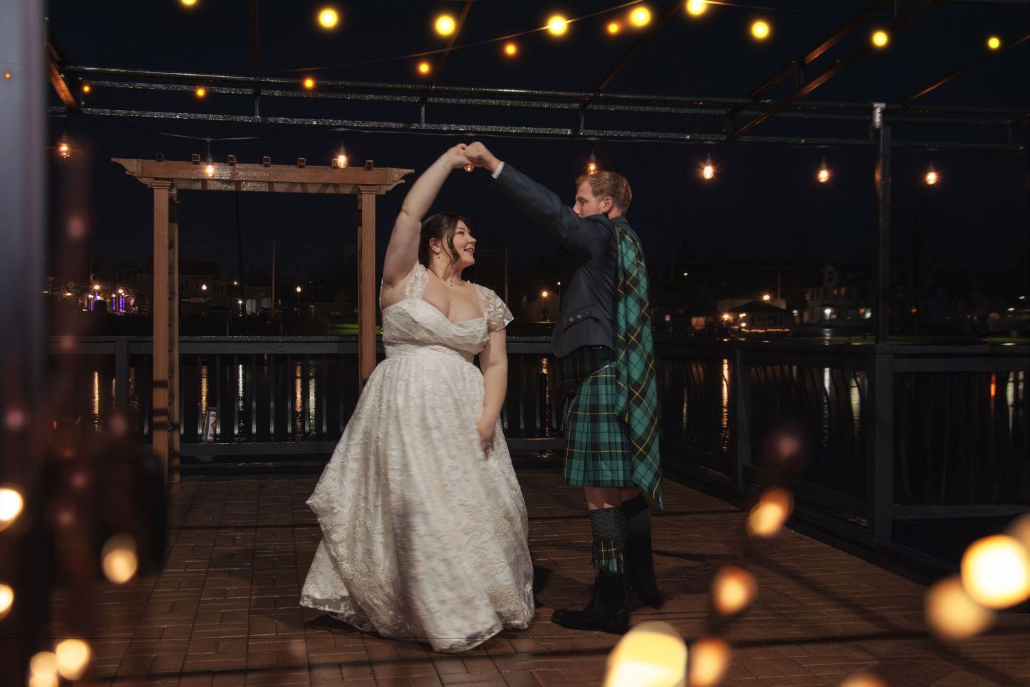 Bride and groom share first dance on outdoor deck at night, surrounded by warm string lights near waterfront.