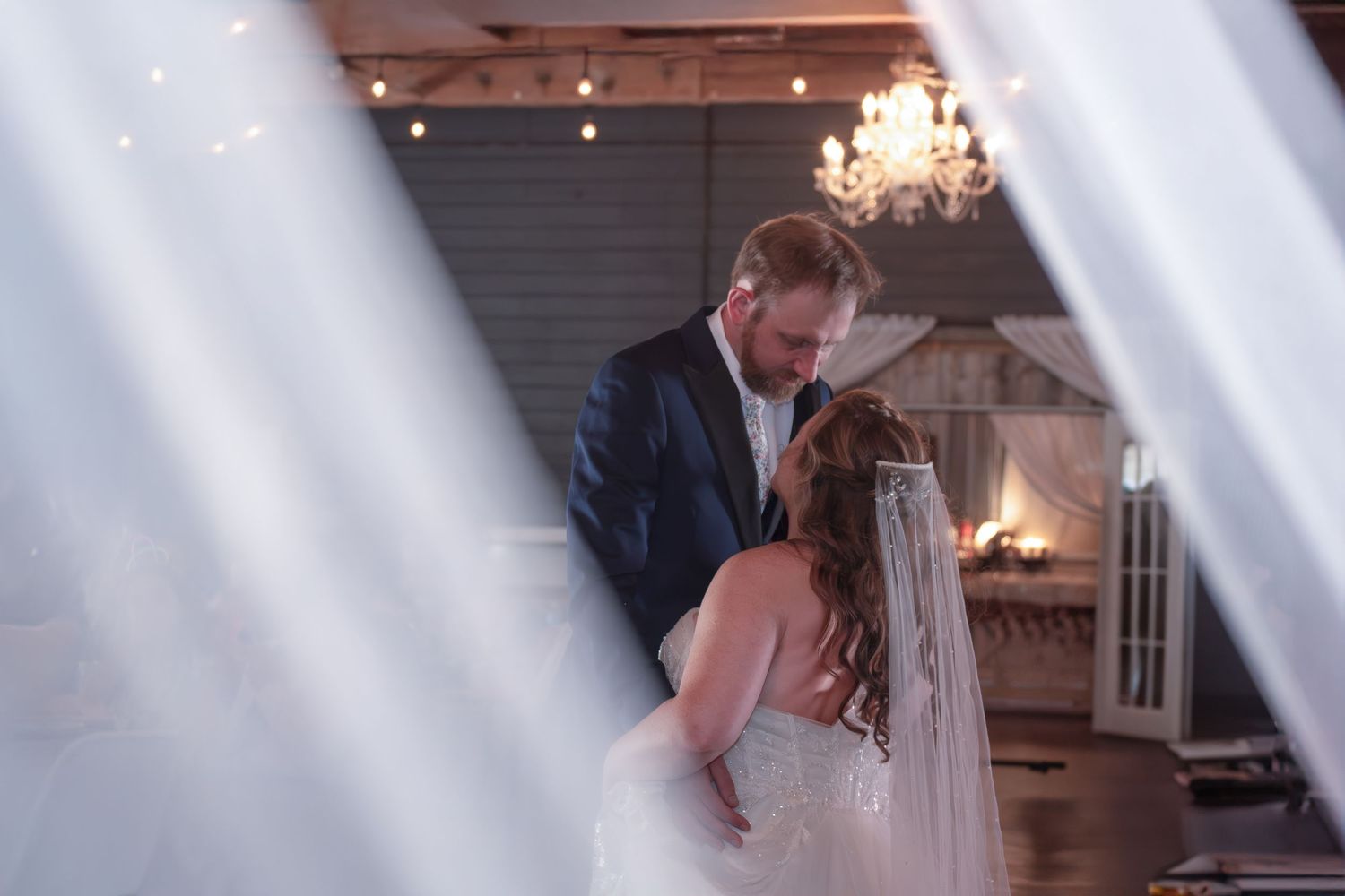 Bride and groom share an intimate moment framed by sheer white curtains, with chandeliers and fireplace glowing behind them.
