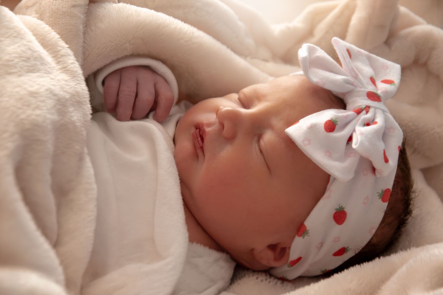 Peaceful newborn baby sleeping wrapped in soft white blanket wearing a polka dot bow headband.