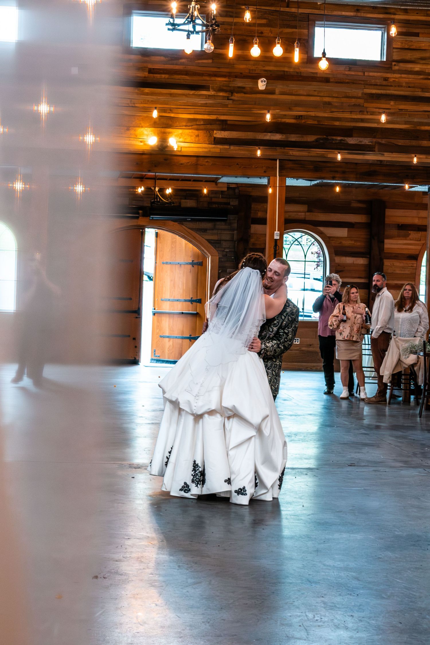 Bride and groom share first dance in rustic barn wedding venue with warm string lights and wood walls.
