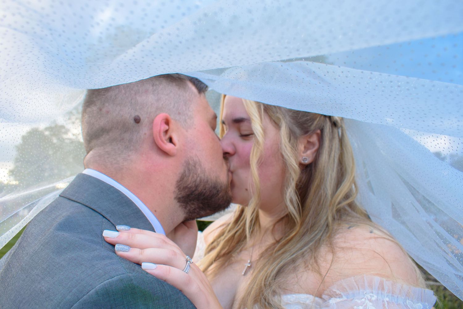 Bride and groom sharing a romantic kiss under a flowing white wedding veil outdoors on their wedding day.
