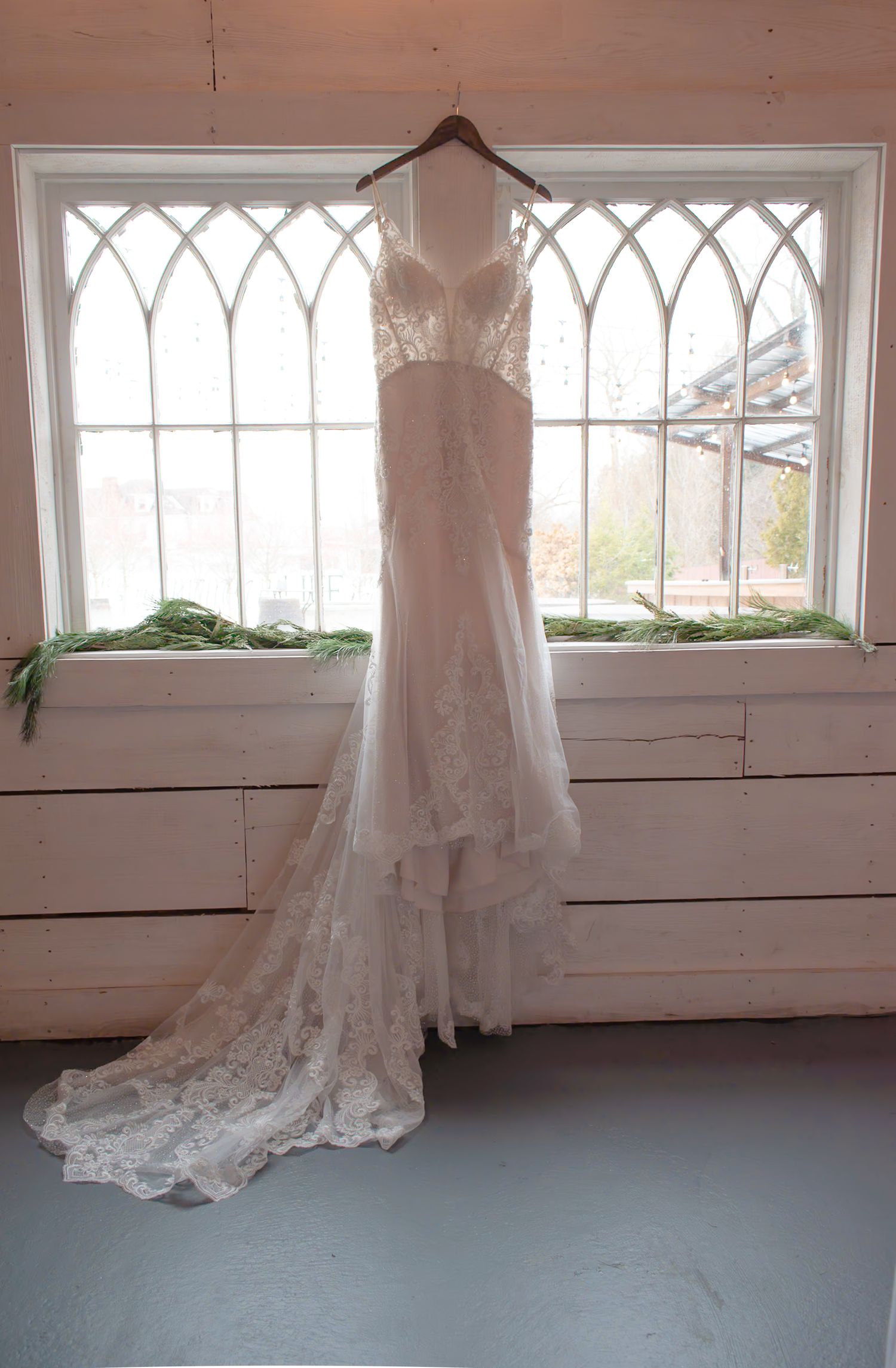 Elegant lace wedding dress with train hanging in a window with gothic arch details and greenery.