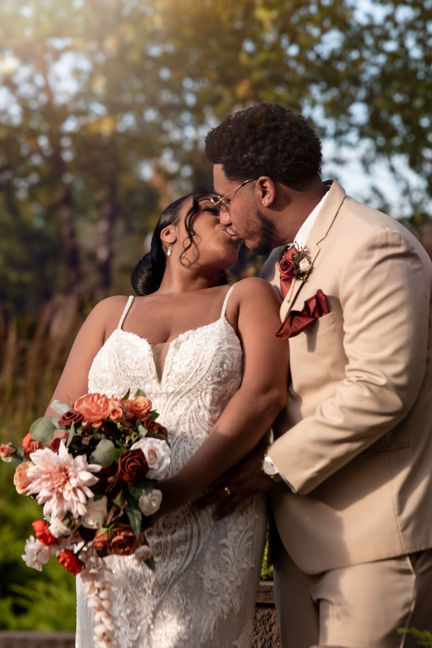 A Black bride and groom share a romantic moment outdoors, she holds a bouquet of red and pink flowers.