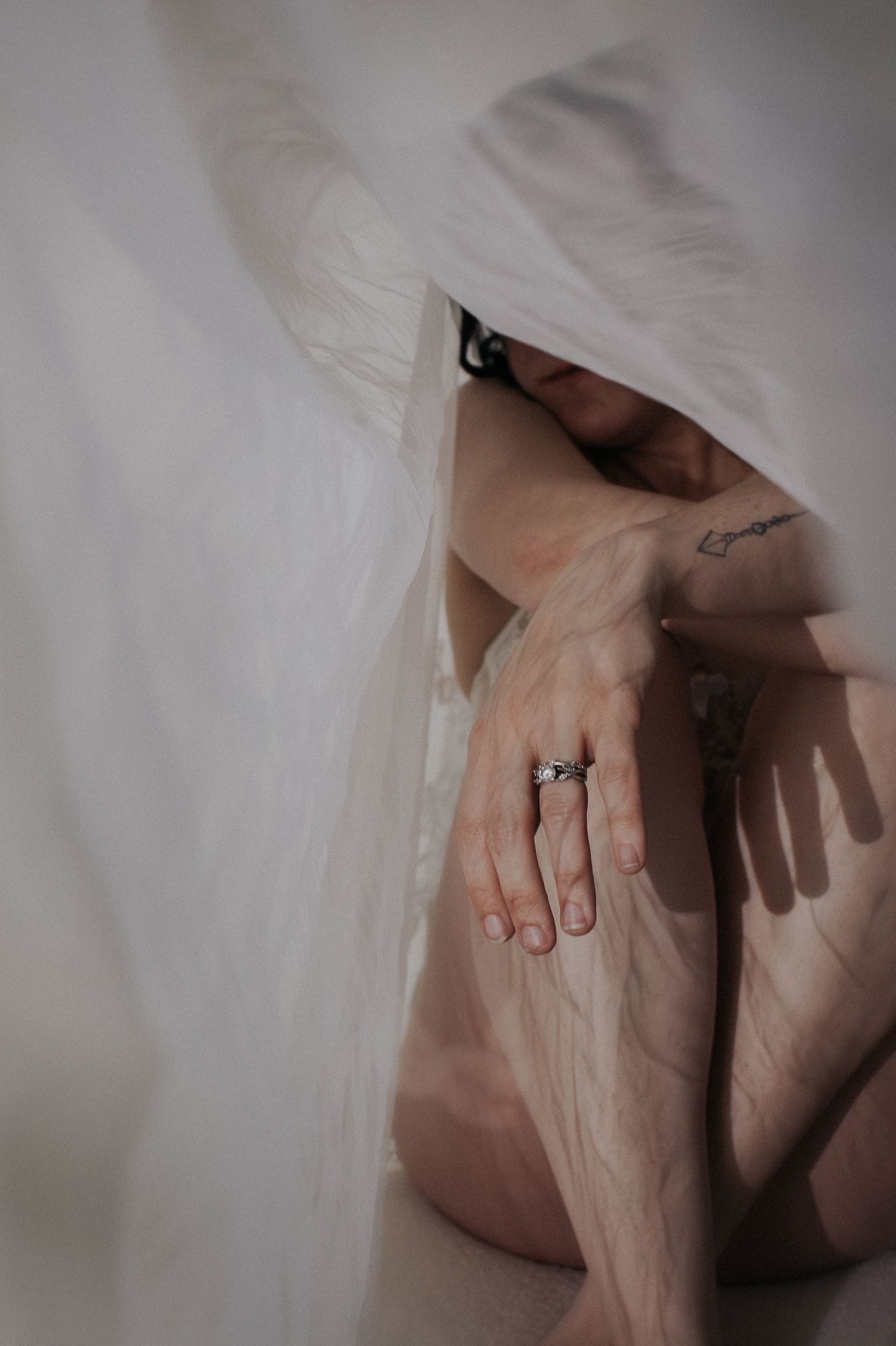 Person draped in sheer white fabric, showing tattooed skin and silver ring, in artistic moody photography.