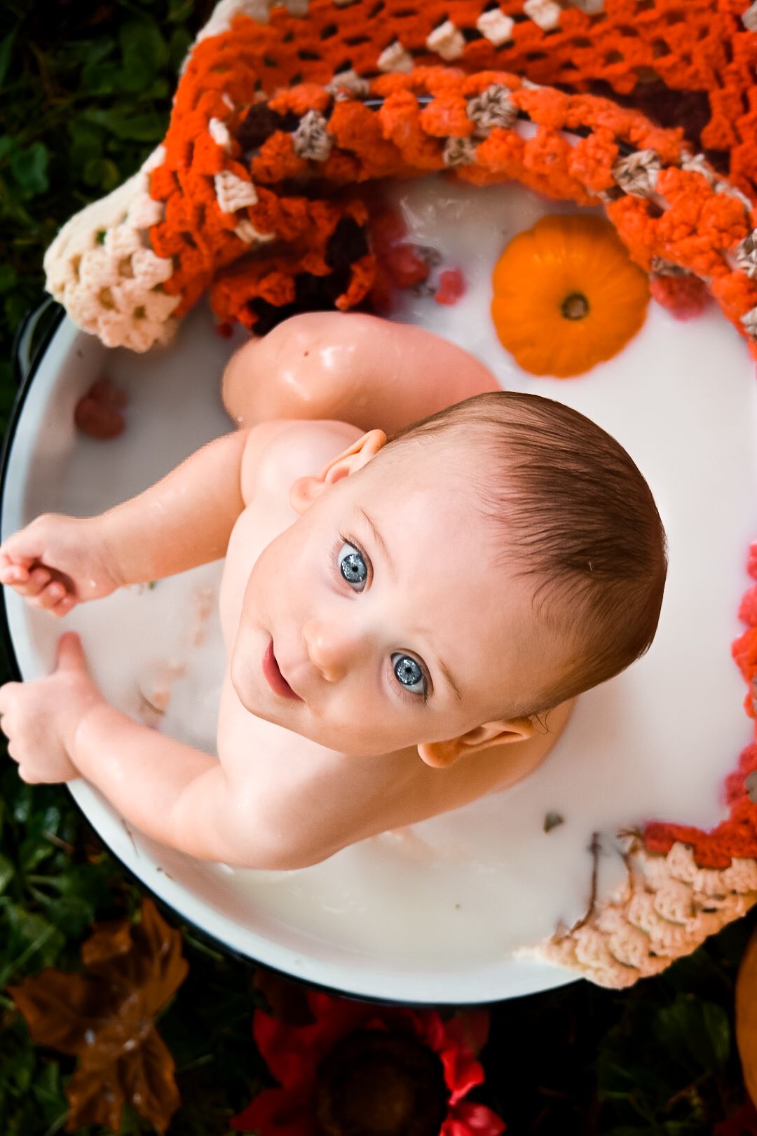 Blue-eyed baby sitting in a milk bath tub outdoors, surrounded by orange crocheted blankets and mini pumpkins.