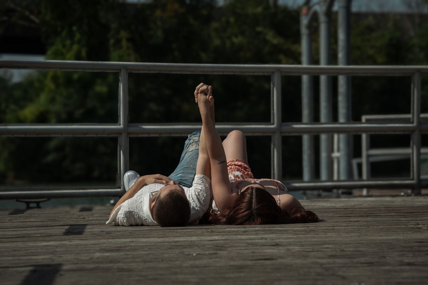 Two people lying on a wooden deck in the sun, relaxing outdoors near a metal railing with trees in the background.