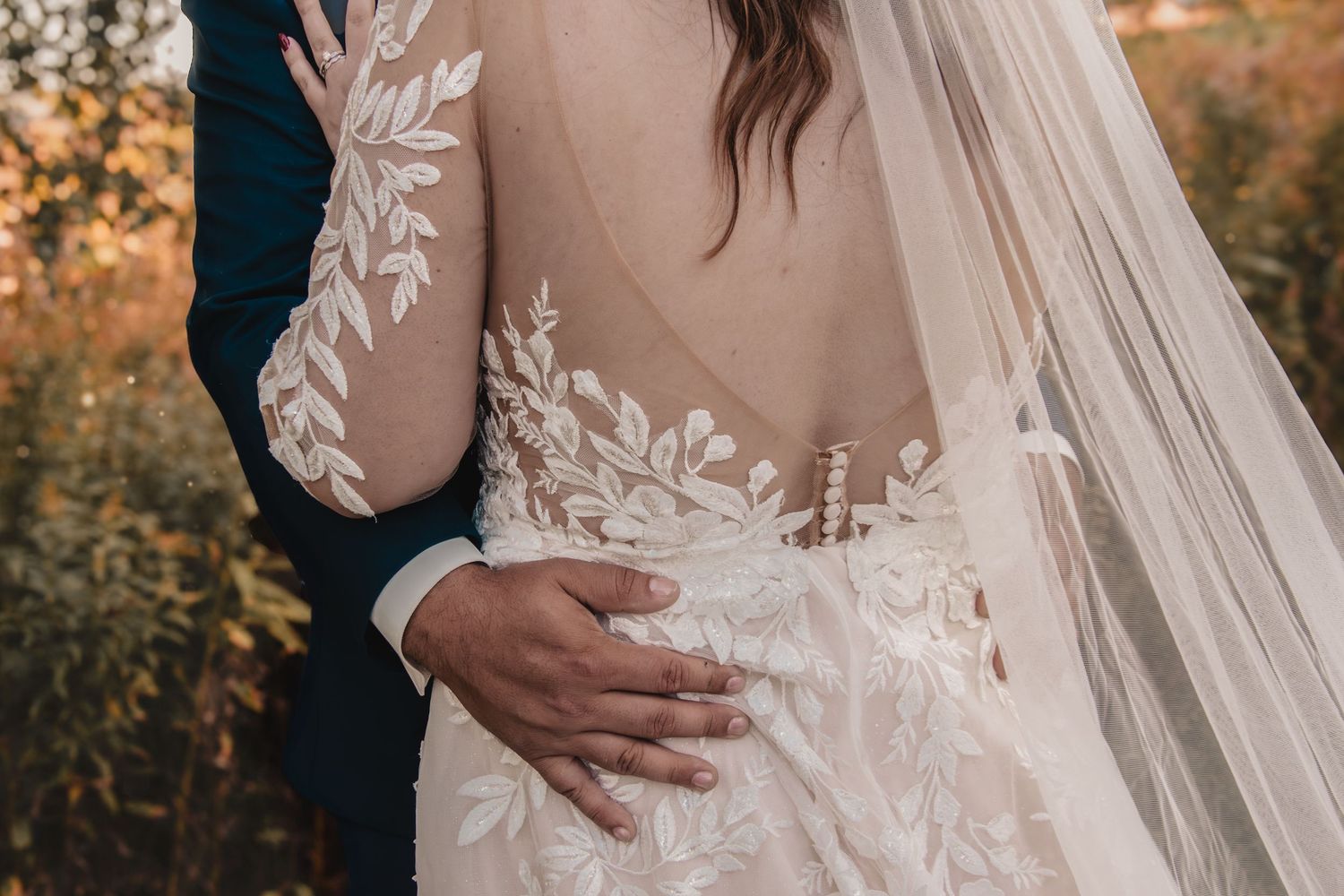 Close-up of a bride in an elegant lace wedding gown with floral embroidery and sheer back, with groom's hand on her waist.
