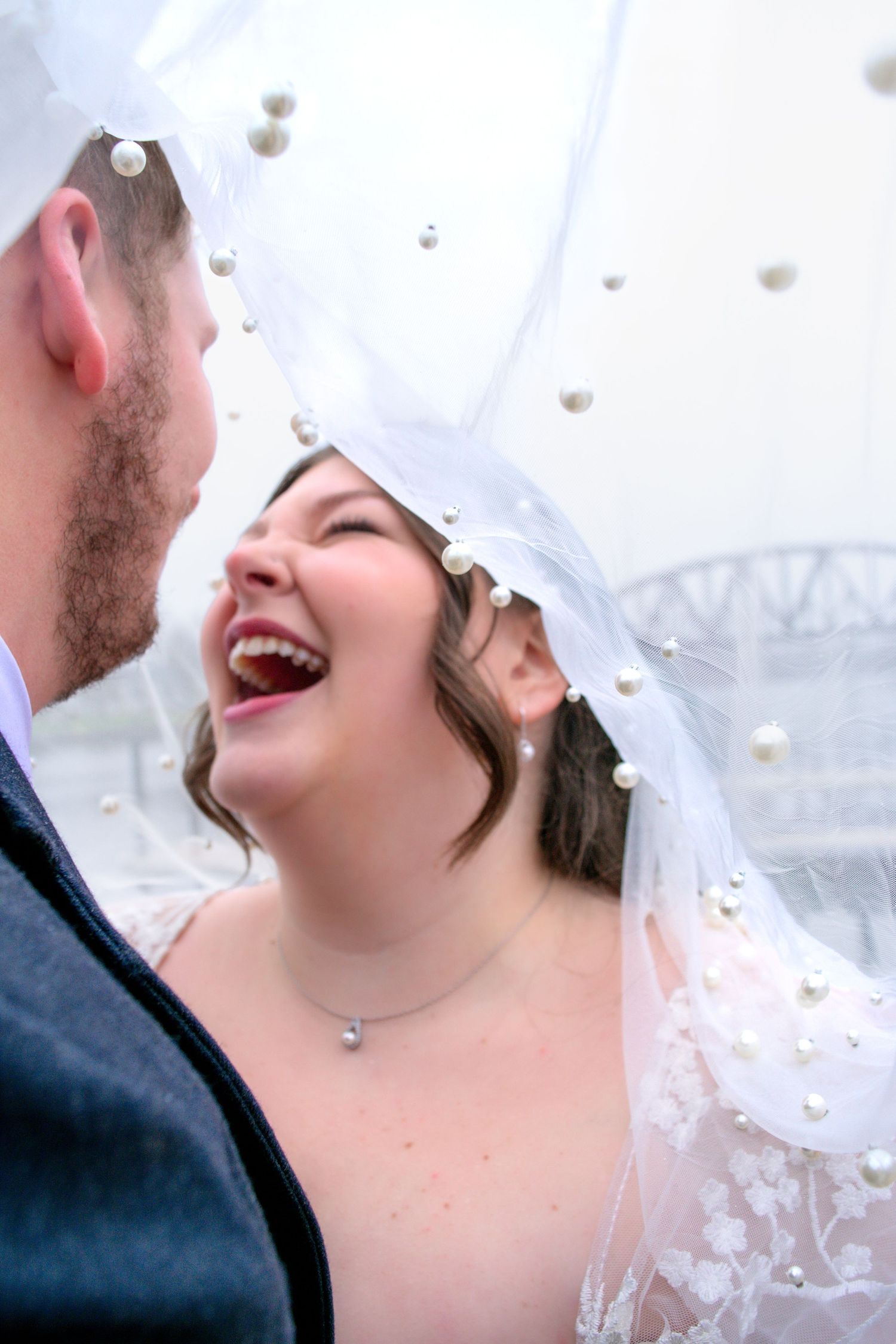 Bride laughing joyfully with groom under pearl-adorned white veil on wedding day.