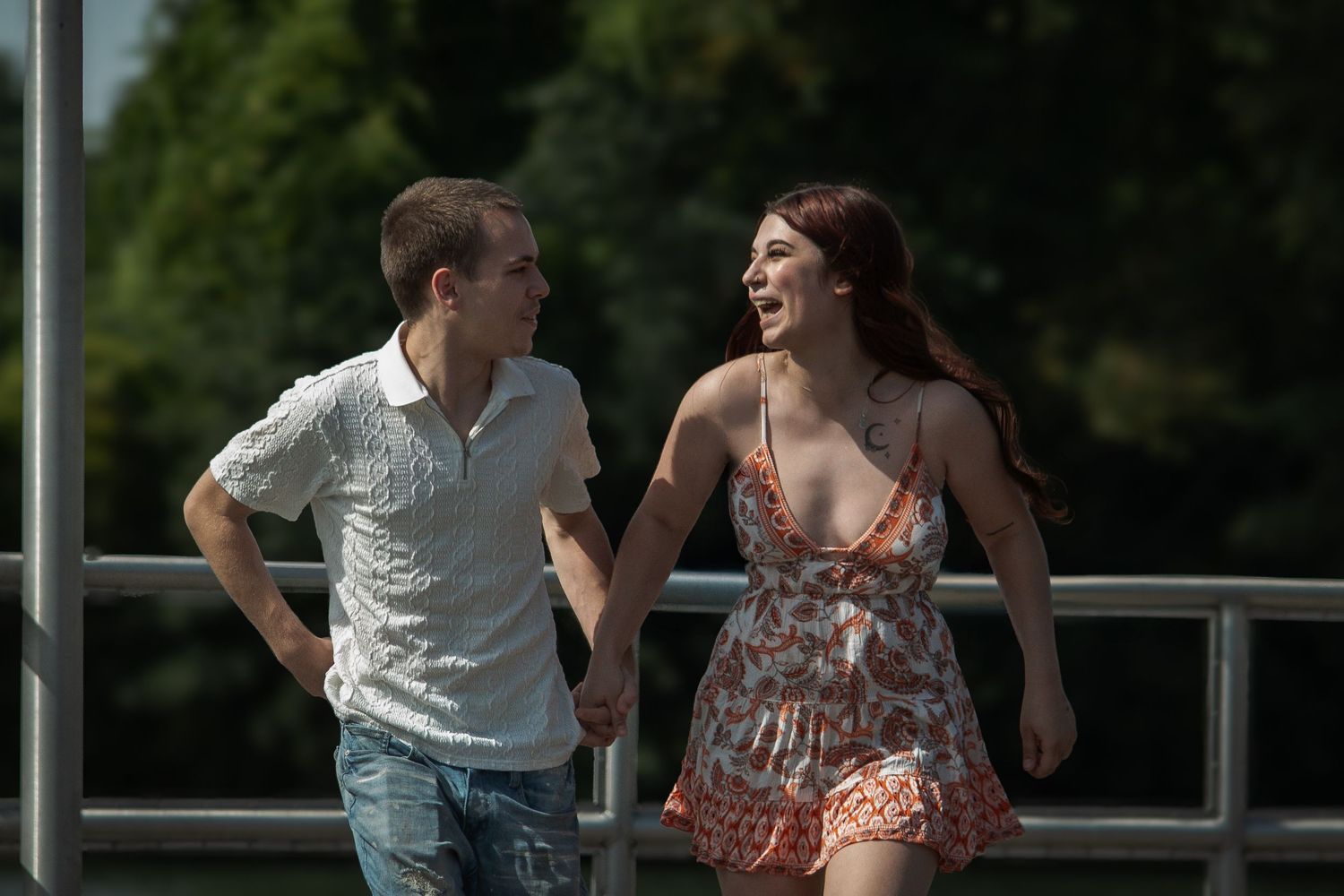 A young couple holds hands and smiles while walking together on a bridge on a sunny day.