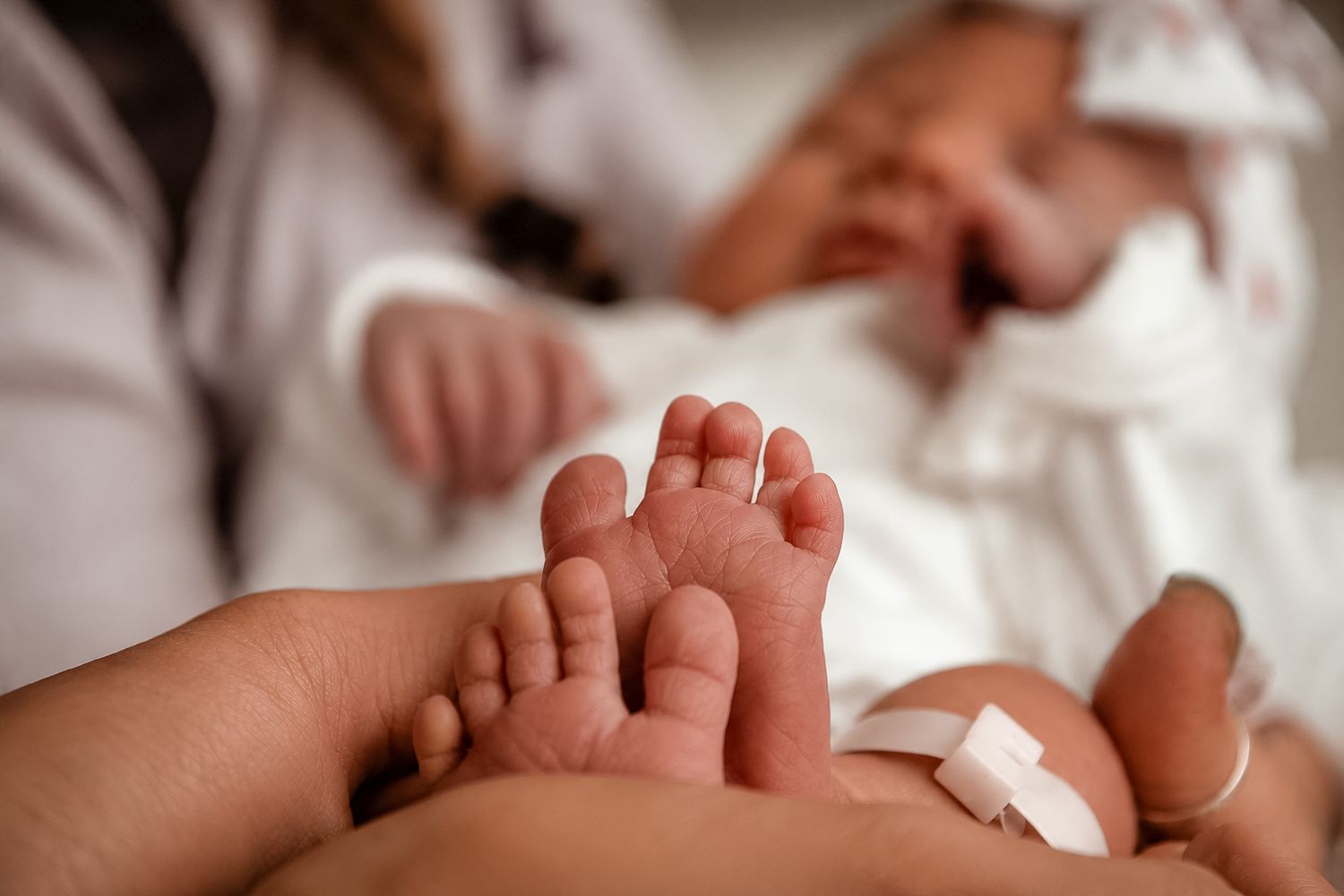 Close-up of newborn baby feet held gently in adult hands, with two infants resting in white blankets in background.