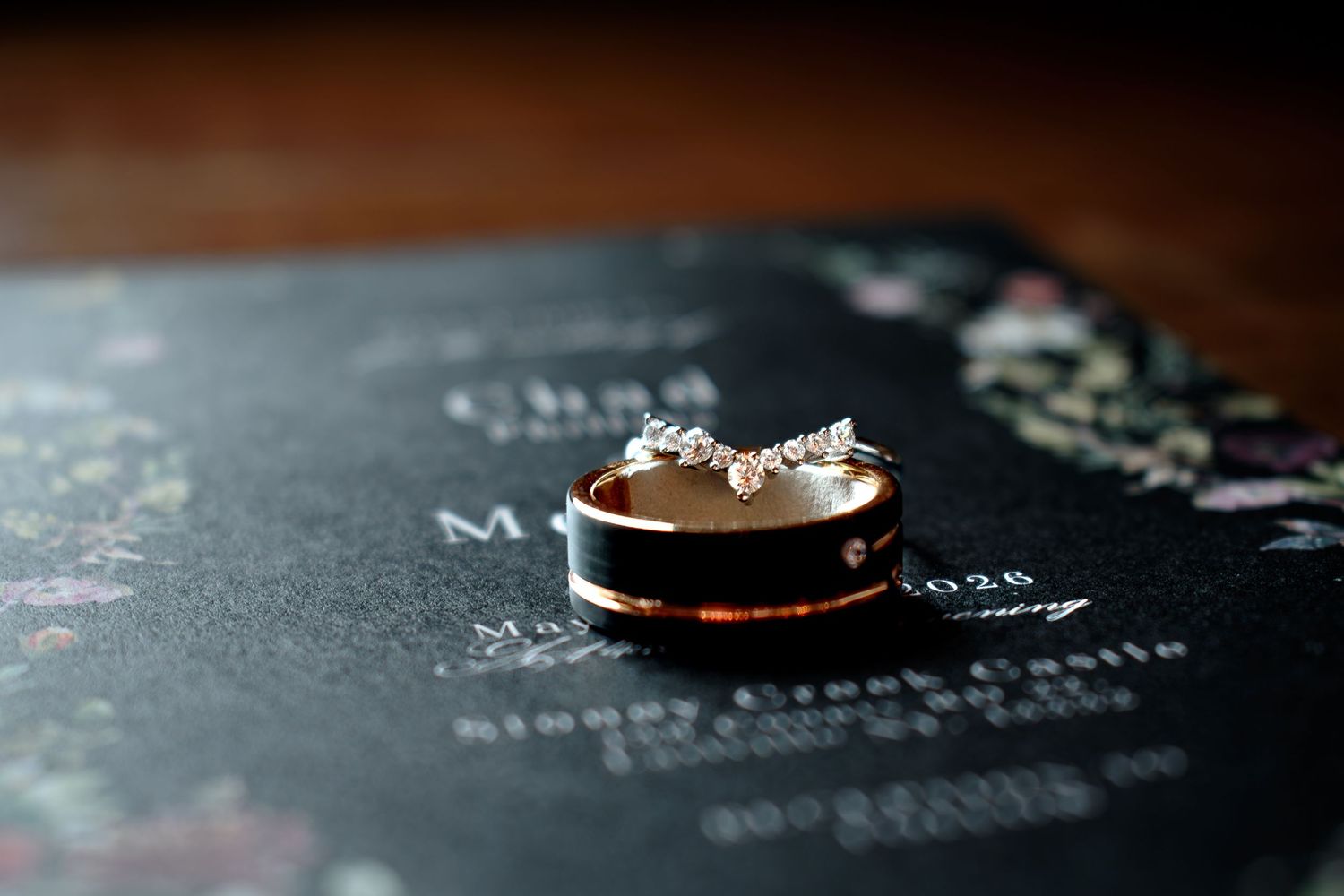 Gold and rose gold wedding rings resting on a dark floral wedding invitation, with a diamond engagement ring balanced on top.