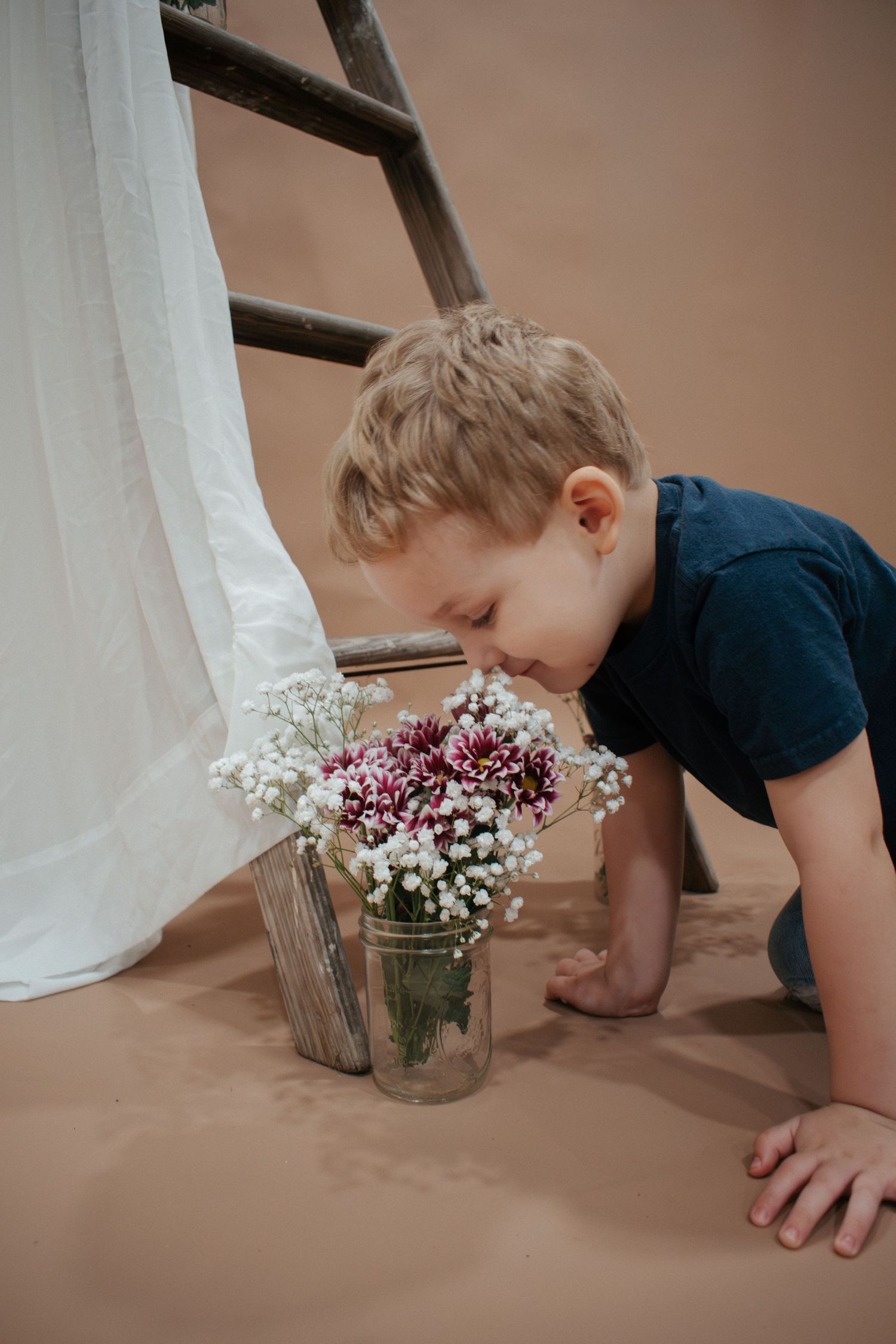 Young boy leaning down to smell colorful flowers in a glass jar near a rustic ladder and white curtain.