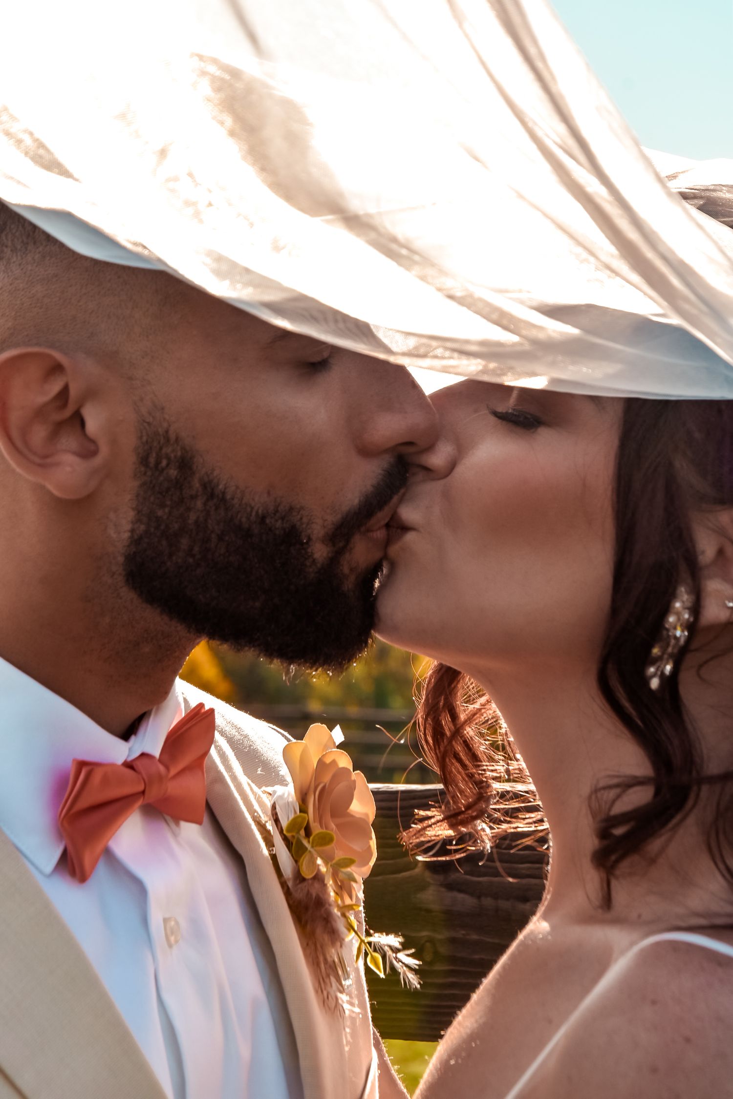 Bride and groom sharing a passionate kiss under a flowing veil with golden sunlight and a lake backdrop.